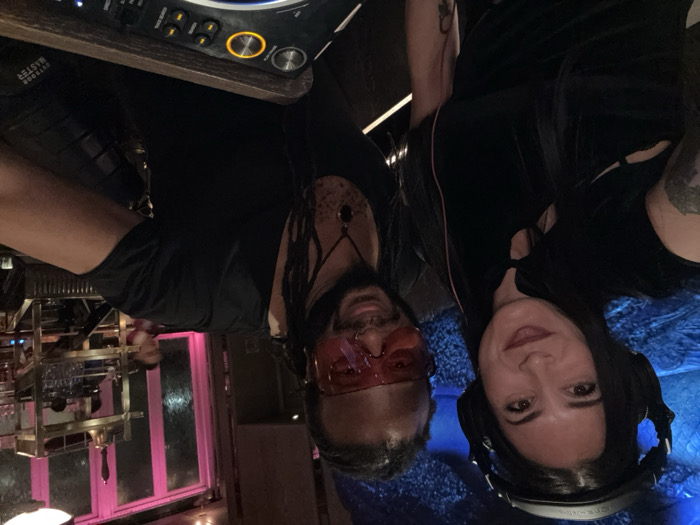 Two individuals, a female and a male, are captured in a close-up photograph within an indoor establishment in London, United Kingdom. The female, positioned on the left, has long dark hair, wears black Audio-Technica over-ear headphones, and a dark, short-sleeved top with a Mandarin collar and a keyhole cutout. A tattoo is visible on her left forearm. She looks directly at the camera with a subtle smile. The male, to her right, has dreadlocks, wears red-tinted sunglasses, a black V-neck top, and a necklace with a dark pendant; a nose ring is visible on his left nostril. He also smiles towards the camera.
The background features a blue, textured wall surface behind the female and a prominent window or glass partition illuminated with pink light behind the male. Through this pink-lit area, another person is dimly visible. A multi-tiered, brass-toned shelving unit holding glassware and a bell is also present in the mid-background. In the lower right foreground, a portion of a professional DJ controller or mixer with illuminated buttons and a wooden-like frame is visible. An object bearing the text "OUTDOOR MASTER" is situated behind the controller. The overall environment is characterized by dim, atmospheric lighting, suggesting a nightlife venue such as a bar or club.