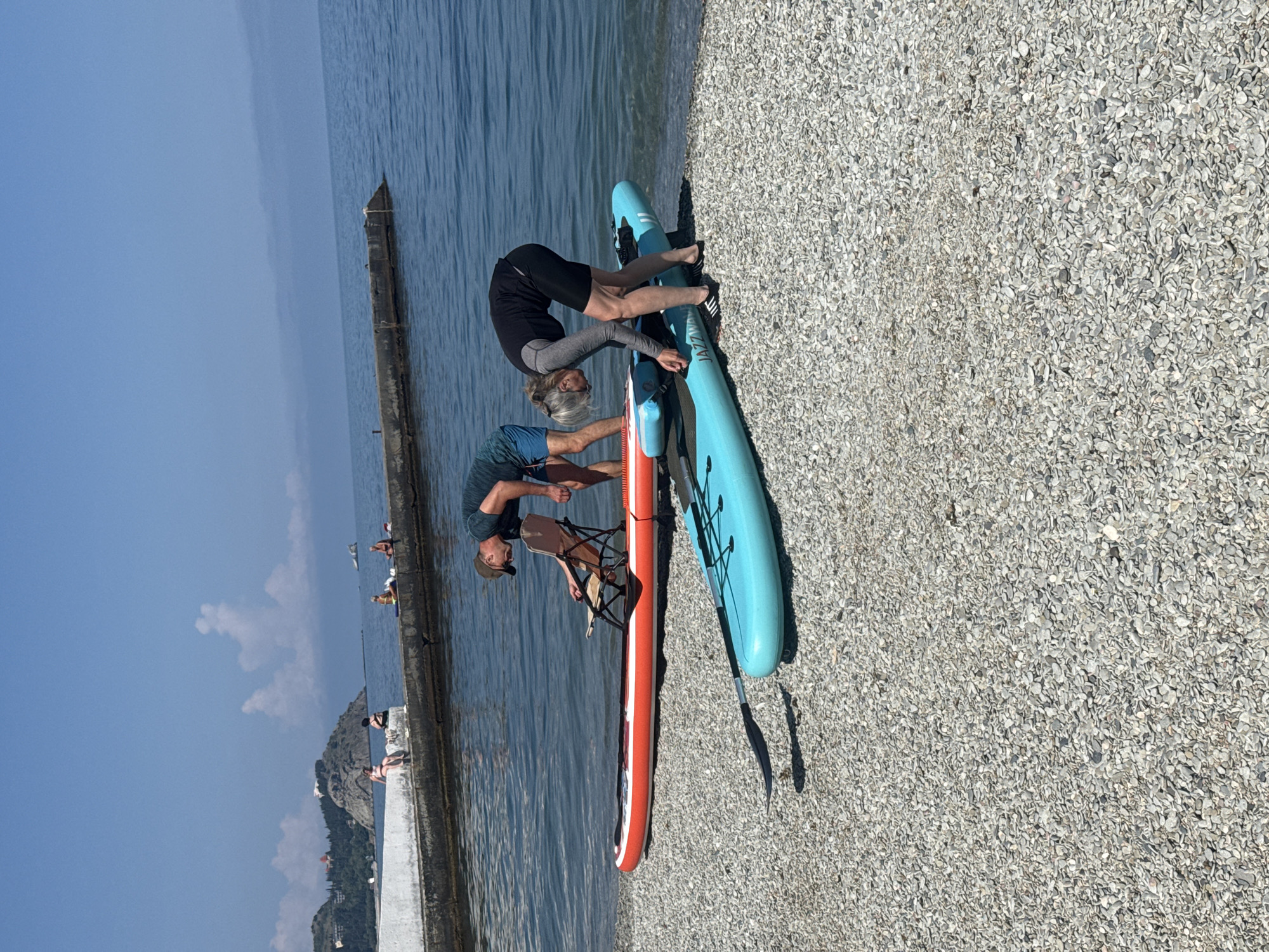 The image shows two individuals on a pebble beach, potentially preparing for water activities. One individual, dressed in dark shorts and a grey long-sleeved shirt, is bending over near a light blue paddleboard. The other individual, wearing a blue shirt and a hat, is also bending over near an orange paddleboard with a folding chair attached. The paddleboards are lying on the pebble beach. In the background, there is a body of water, possibly a sea or lake, with a concrete pier or structure extending into it. Other people are visible on the pier. The location is Unknown, Unknown.