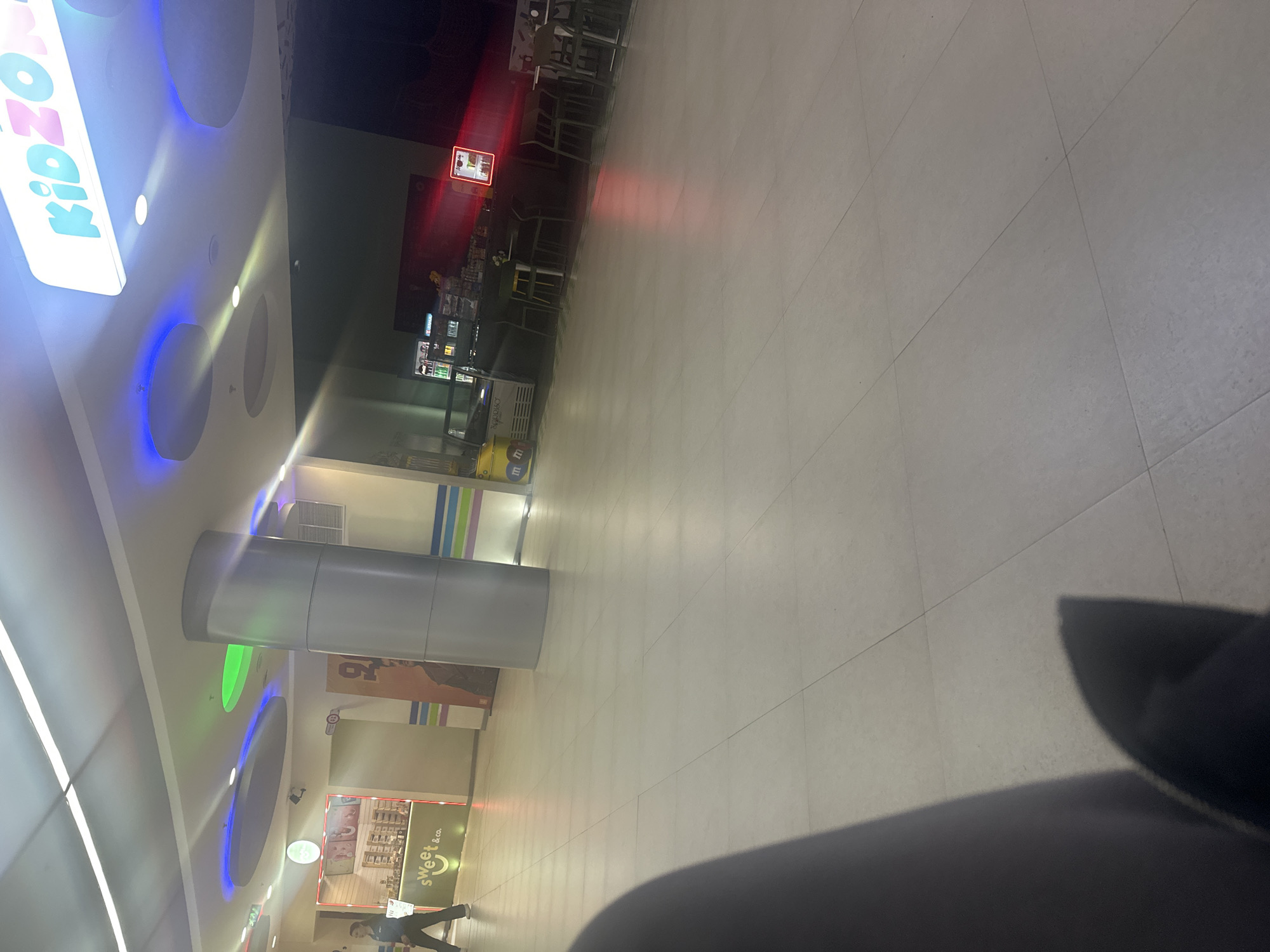 The image shows an indoor shot of a shopping mall in Yerevan, Armenia. A large, cylindrical pillar sits in the center of the frame. In the top left, a "KIDZONE" sign is visible, while at the bottom is a "Sweet & Co." store. A person can be seen walking near the bottom of the image. The floor consists of large, square tiles. In the upper-left corner, there are tables and chairs, possibly belonging to a food court area.