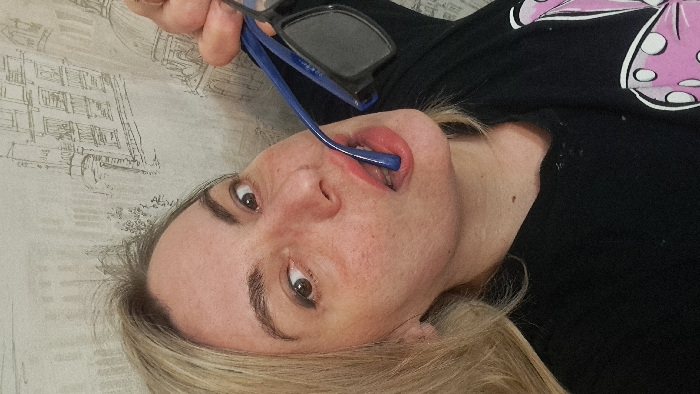 An indoor scene in Bel'kovo, Russia, features a blonde-haired woman with fair skin, brown eyes, and dark eyebrows, directly facing the camera with a wide-eyed expression. She wears a black v-neck shirt featuring a pink and white graphic on the lower right. Her right hand holds a pair of blue-framed sunglasses with dark lenses, and one arm of the sunglasses is positioned inside her mouth, between her lips. The background is a light-colored wallpaper patterned with monochrome line drawings of architectural structures, including multi-story buildings and domes.