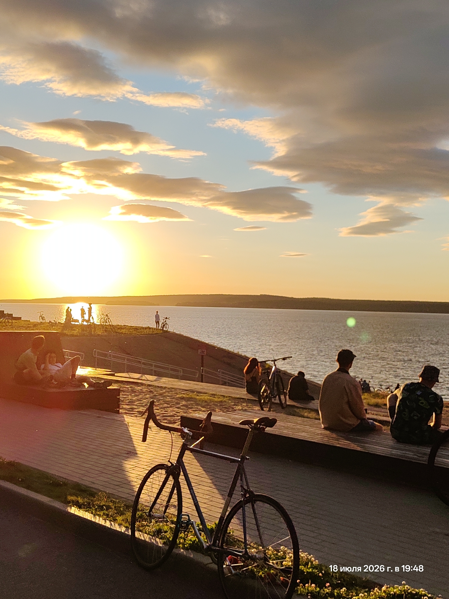 The image captures a serene sunset scene at an unknown location, likely a waterfront park or promenade. The sun, a bright, large orb, is setting over a wide body of water, casting a warm, golden glow across the sky, illuminating scattered clouds with shades of orange and yellow. In the foreground, a paved walkway with scattered greenery lines the edge of the water. Several people are visible, some sitting on benches or the ground, enjoying the view. A bicycle is parked prominently in the lower right quadrant of the image. Further down the walkway, silhouetted figures and bicycles are visible against the setting sun. The overall atmosphere is peaceful and relaxed, with a sense of people enjoying the end of the day. A timestamp in the lower right corner reads "18 июля 2026 г. в 19:48," which translates to "July 18, 2026 at 19:48."