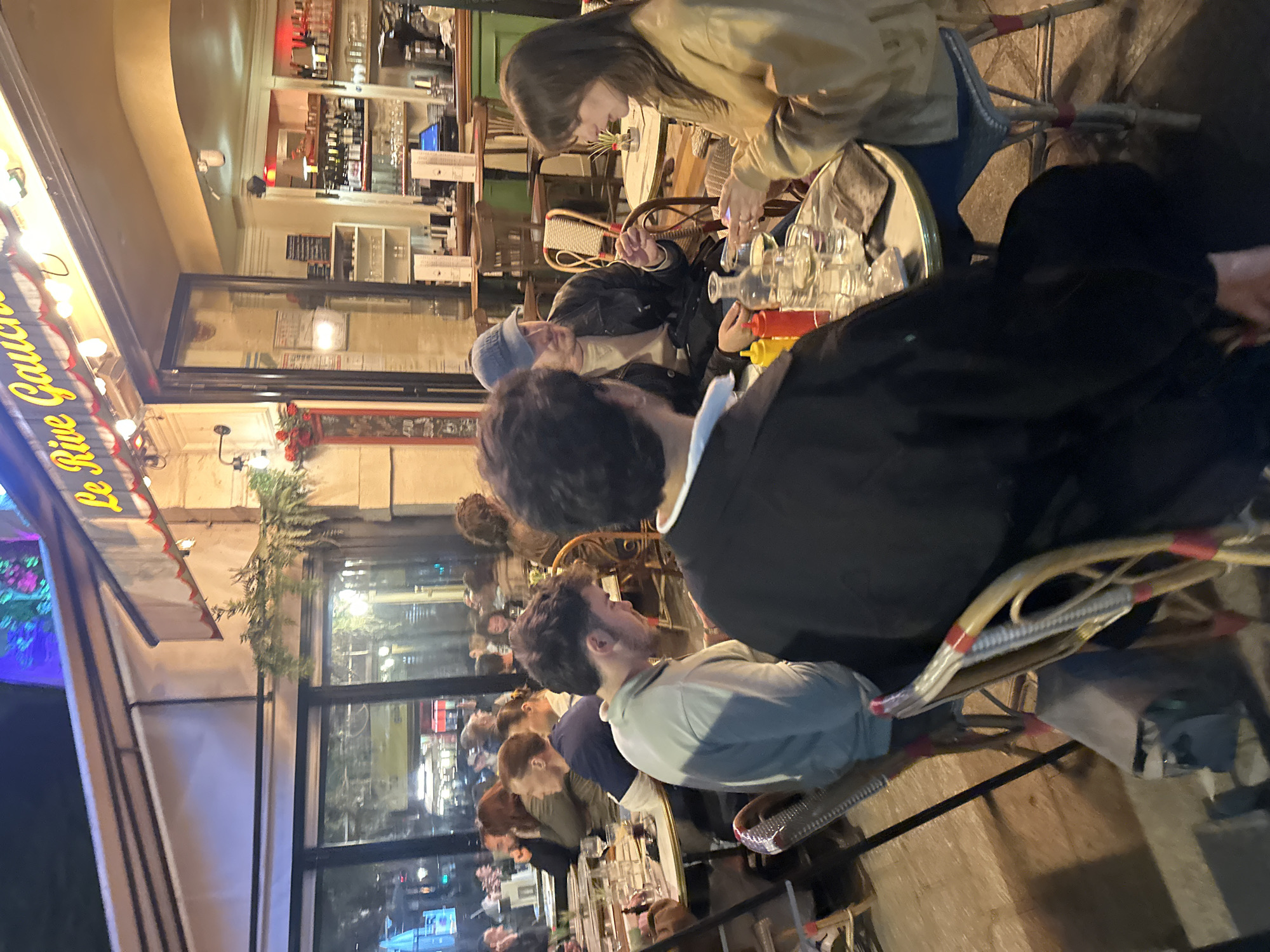 The image depicts a street scene at an outdoor café in Paris, France, specifically "Le Rive Gauche." Several people are seated at tables, some conversing, while others appear to be occupied with phones and tables. The environment is characterized by outdoor seating, visible interiors of the restaurant with bottles and bar, and street views with passing traffic and lights. The time of day is likely evening, judging by the dim lighting and outdoor illumination. The foreground features people at the café tables, and the background shows the streetscape.