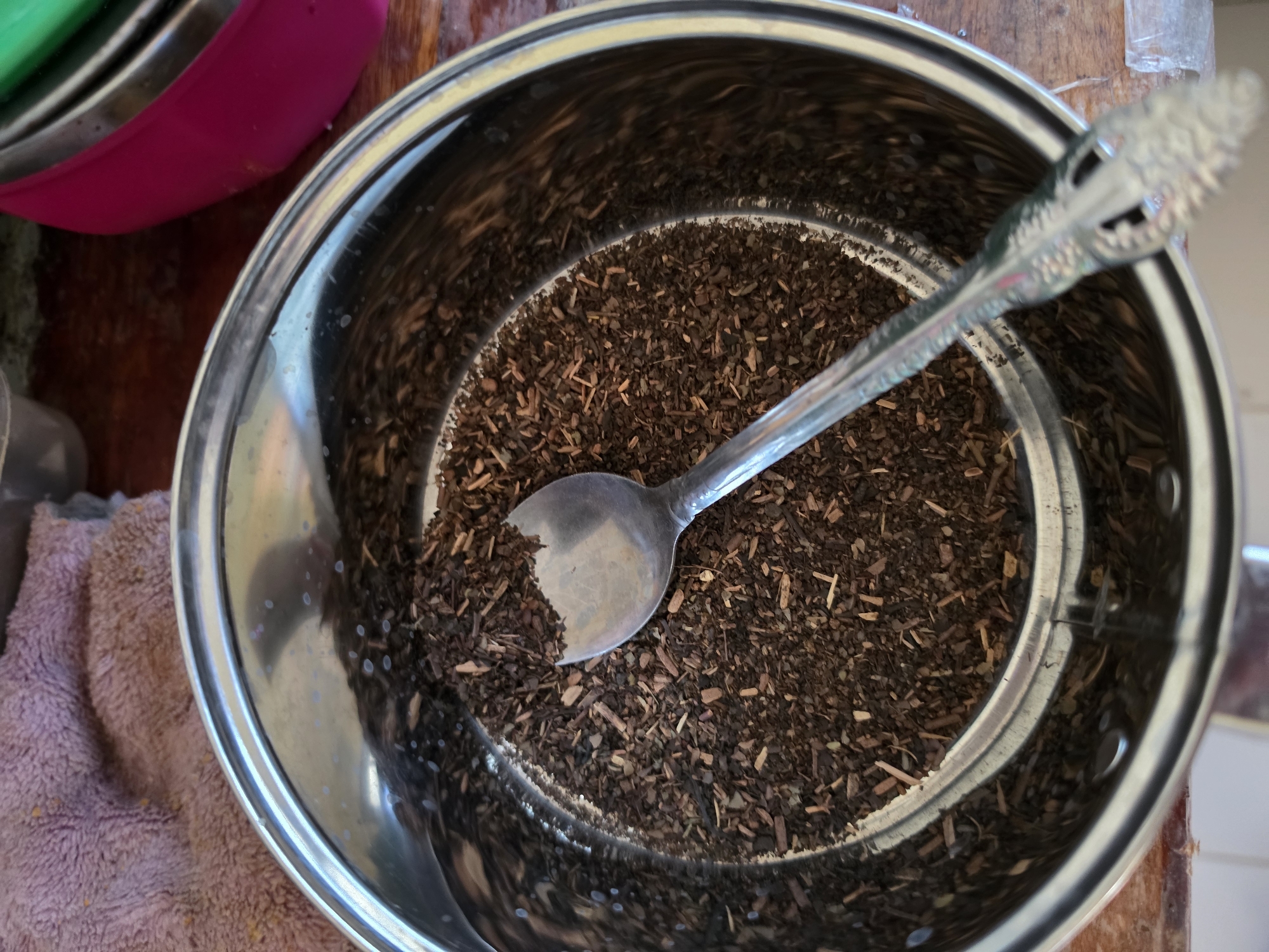 A close-up, top-down view shows a metal pot filled with loose-leaf tea. A silver spoon rests in the tea, with its handle extending out of the pot. The pot is on a wooden surface. In the background, a pink towel and part of a pink bowl are visible. The scene appears to be indoors.