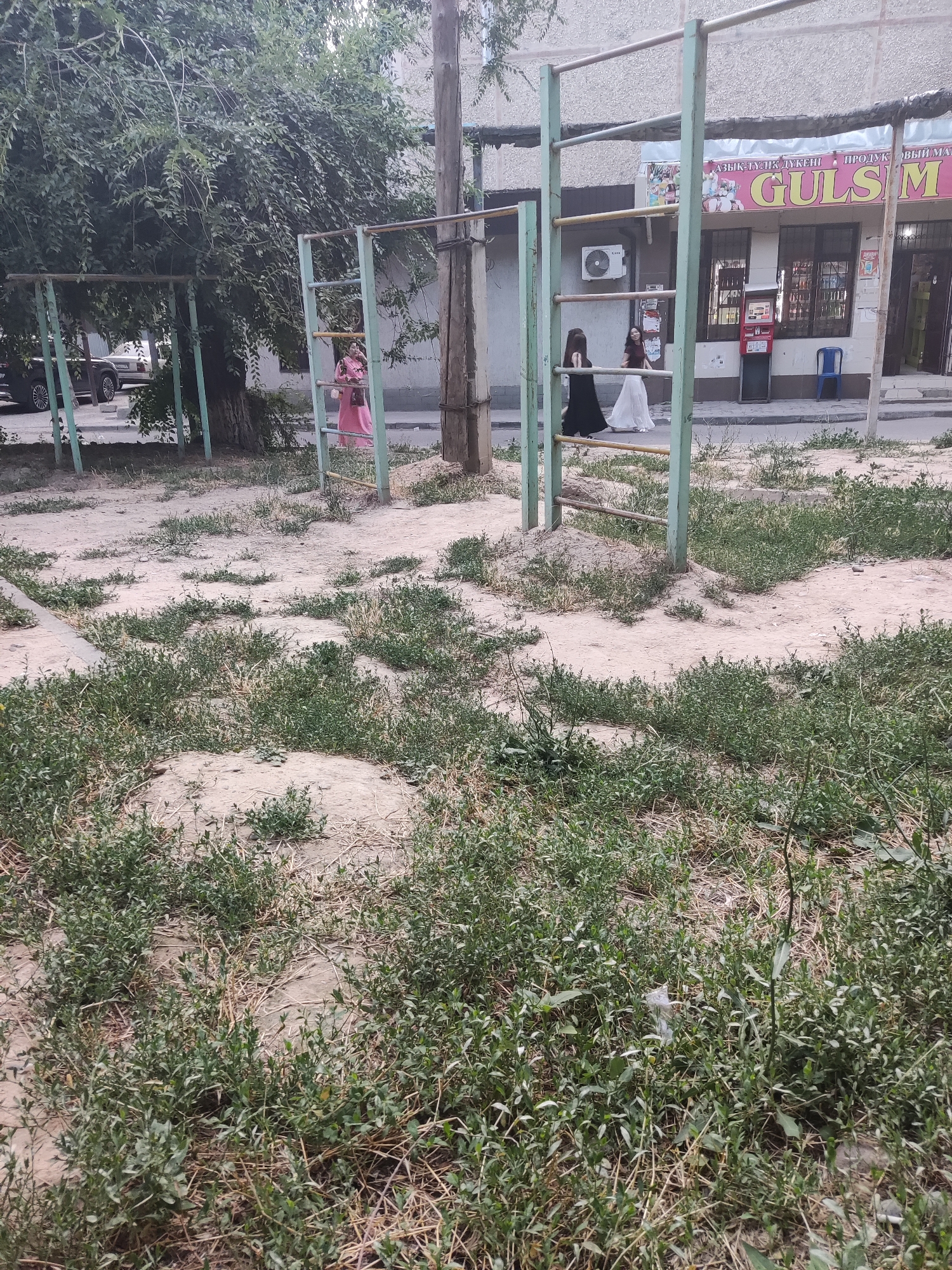 The image depicts an outdoor scene in a city, likely Shymkent, Kazakhstan, featuring a public sports ground with horizontal bars and pull-up bars. In the background, a storefront with a large sign reading "GULSM" is visible, suggesting a commercial establishment. Three individuals are present: one person in a pink dress stands near the sports equipment, and two individuals in dresses, one black and one white, are walking further down the street. The ground is a mixture of sand and sparse vegetation, indicating a somewhat unkempt public area. A tree with dense foliage is on the left side of the image, and a utility pole is present on the right.