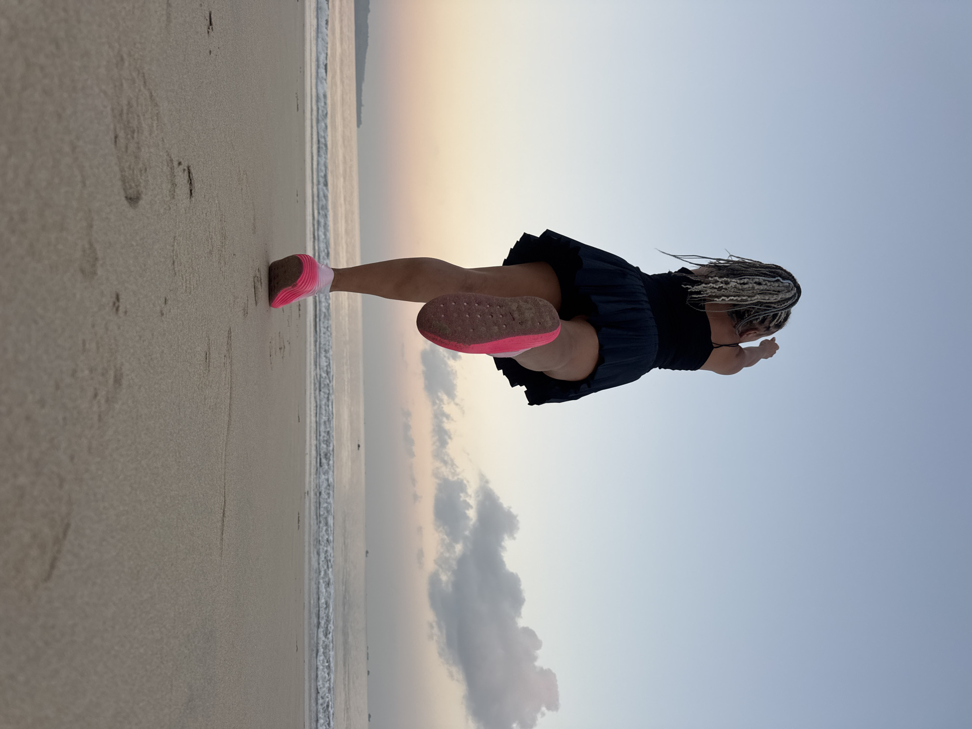 A woman with blonde dreadlocks runs along a sandy beach in Kuta, Indonesia, with the ocean and a pastel sky in the background. She wears a black tank top and a black pleated skirt, along with bright pink running shoes. The camera angle is low, emphasizing the footprints in the sand and the woman's stride. The sun is low on the horizon, casting a warm glow on the scene, and a few boats are visible in the distance on the water.