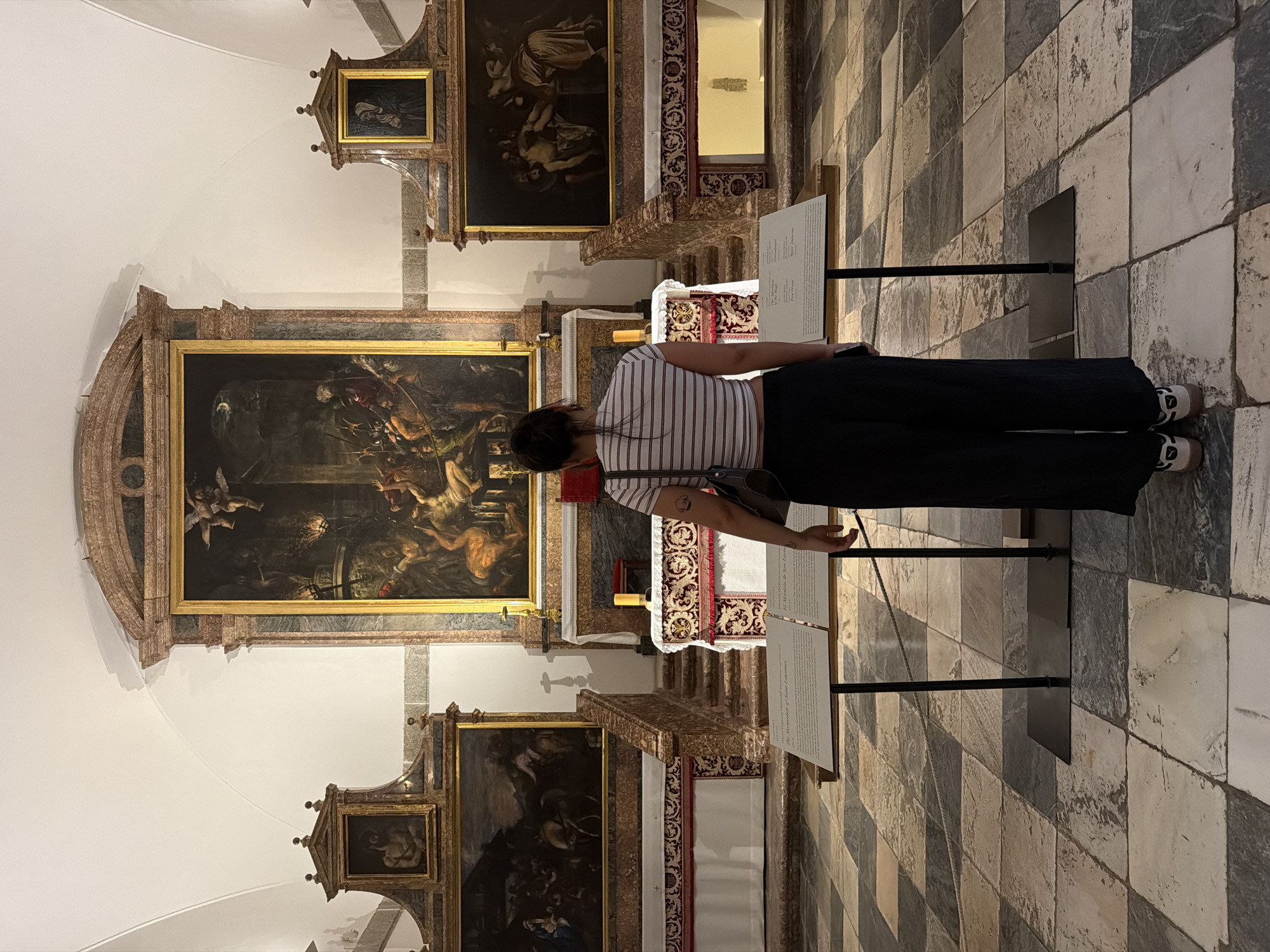 A person stands in a dimly lit, opulent room, likely a chapel or museum exhibit, at San Lorenzo de El Escorial, Spain. The person, seen from the back, is wearing a striped t-shirt and dark trousers, looking at a large, framed oil painting that depicts a dramatic, possibly biblical scene with multiple figures and an angelic presence. The painting is ornate, set within a gilded frame, and is part of a larger display that includes other framed artworks and religious artifacts. The floor is tiled in a checkerboard pattern of light and dark stone. Two display boards with text are positioned in front of the person, suggesting an informational exhibit. The lighting is subdued, with spotlights highlighting the artwork. The atmosphere appears quiet and contemplative. The visible text on the display board mentions "The Martyrdom of Saint Lawrence," indicating the subject of the artwork.