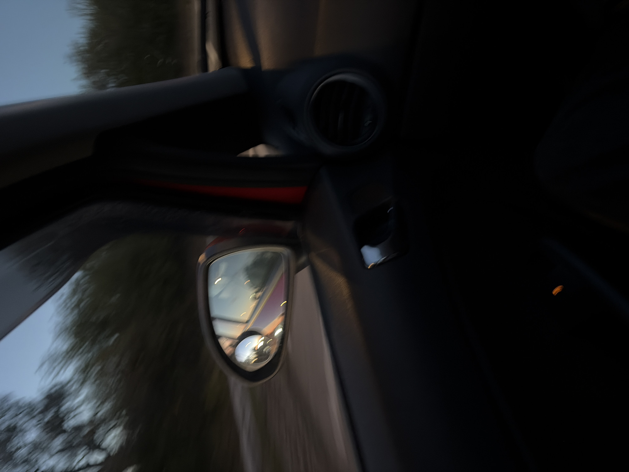 The image shows the interior of a vehicle, likely a car, with a focus on the driver's side mirror and dashboard elements. The mirror reflects a blurred scene of trees and sky, indicating movement. A circular air vent is visible on the dashboard. The surrounding environment is dark, suggesting either nighttime or a low-light condition inside the vehicle. The location in East Suffolk, United Kingdom is not directly visible and would require further information to confirm.