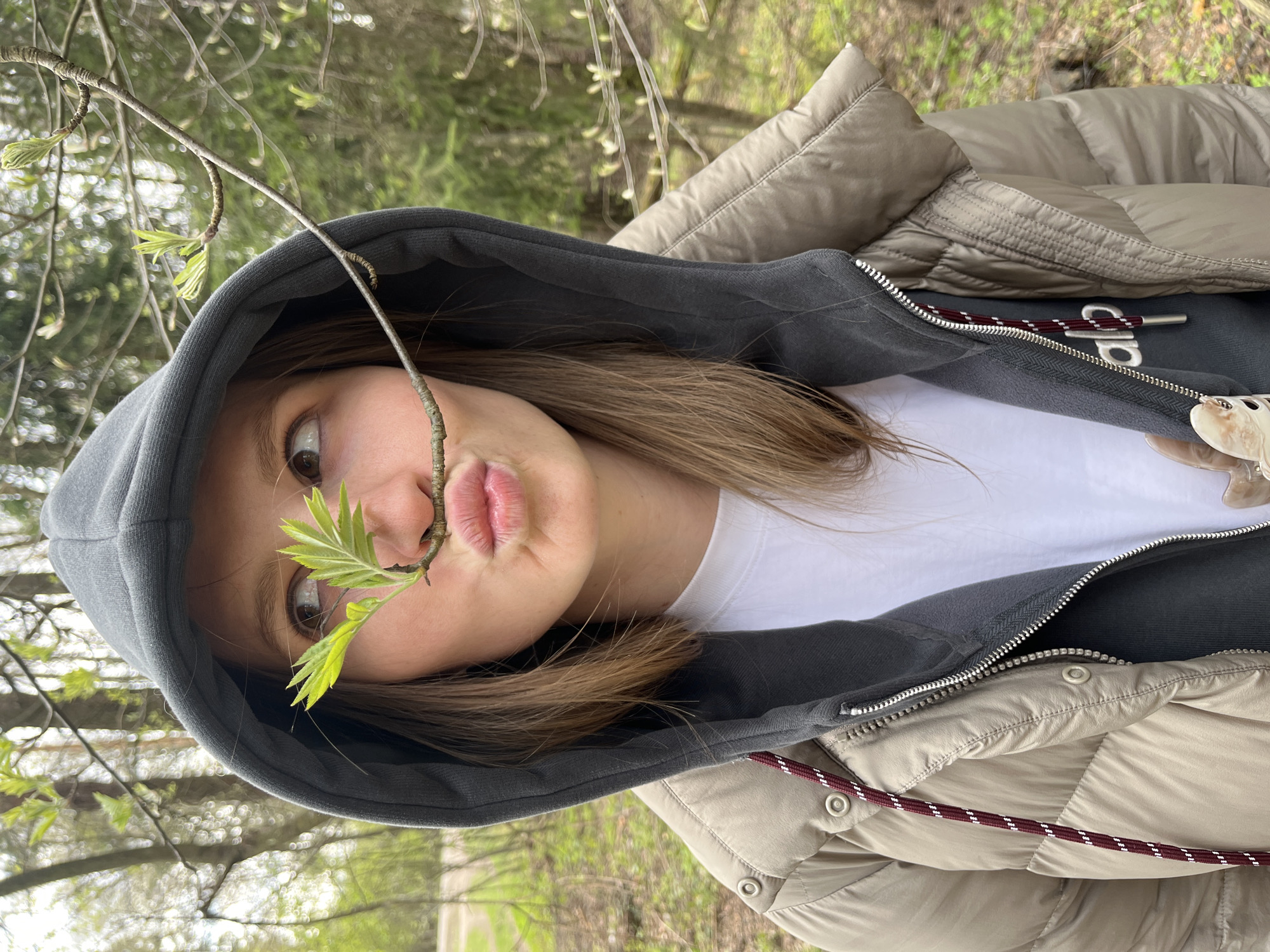 A young woman is centered in the frame, likely photographed outdoors in a wooded area near Razdolyye, Russia. She is wearing a light beige puffer jacket over a gray hooded sweatshirt and a white t-shirt. The hood is up. A thin tree branch with emerging green leaves extends across her face, between her nose and puckered lips. The background shows blurry trees and foliage suggesting a forest setting. The orientation of the photo suggests it was taken at an odd angle, possibly tilting the phone.