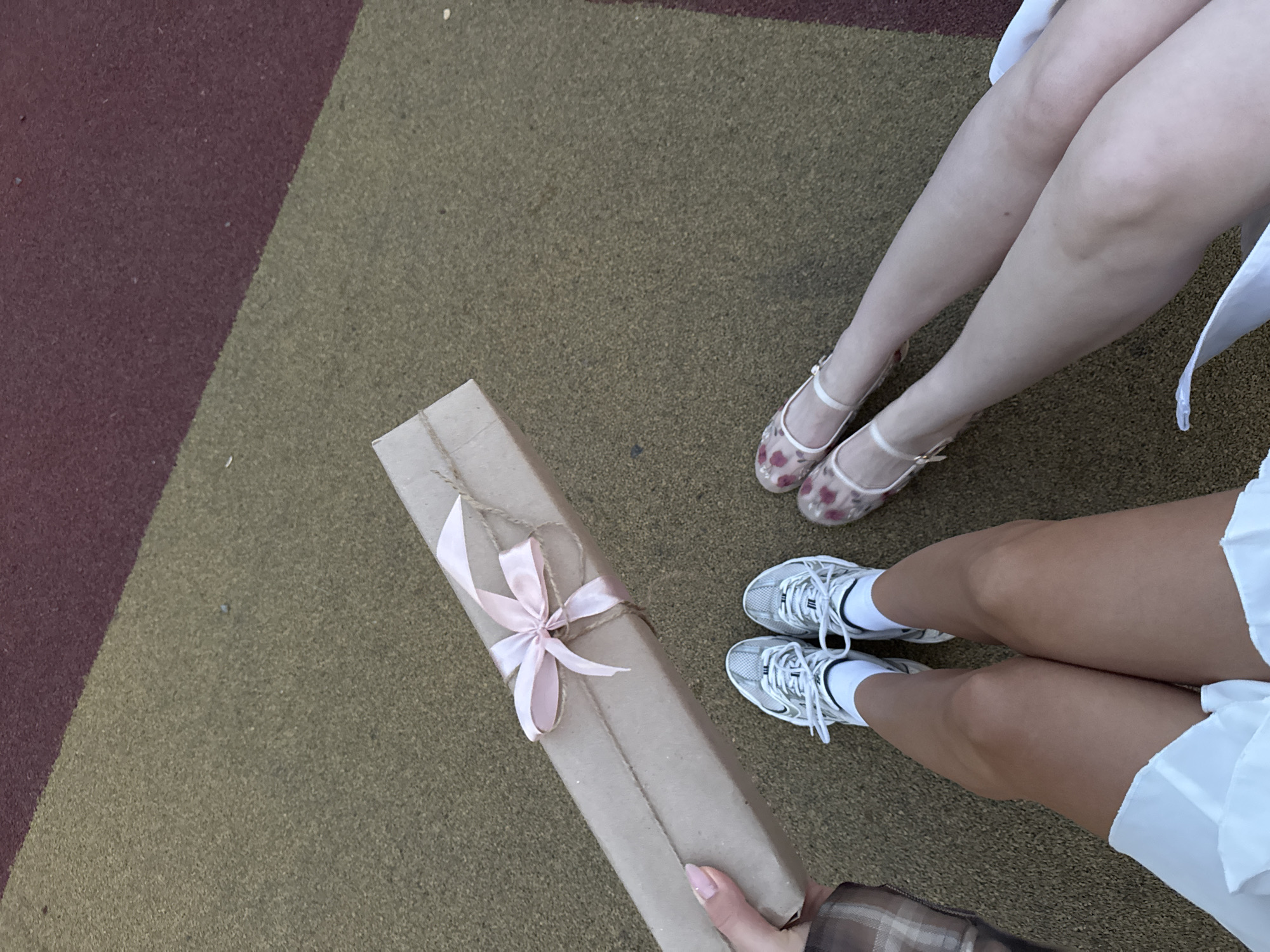 The photograph shows a top-down view of two individuals' legs and a long, thin gift wrapped in brown paper and tied with a pink ribbon and twine. The foreground includes a hand holding one end of the gift. One person has light-colored legs and is wearing floral-patterned shoes with white straps. The other person is wearing white sneakers with white socks. The legs are positioned next to each other. The background shows a two-toned surface, with one area being a dark, reddish-brown and the other a lighter olive-green color. The setting appears to be outdoors, possibly on a playground surface.
