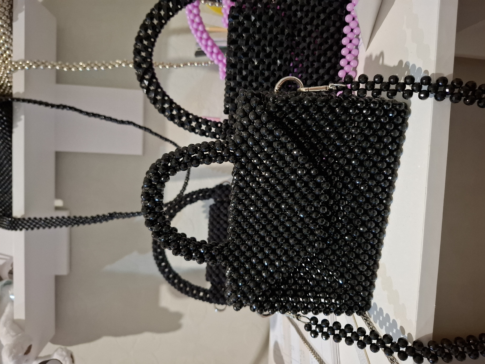 The image shows a close-up, eye-level view of several handmade beaded bags displayed on a white shelf. The most prominent bag in the foreground is a small, rectangular black beaded handbag with a structured shape and short handles. It is made of small, faceted black beads that create a sparkling texture. A detachable beaded strap in a similar black color hangs from the side of the bag. Behind this bag, another black beaded bag is partially visible, also with a handle. Further in the background, there are glimpses of other beaded bags, one appearing to be silver or clear beads, and another with pink beaded elements. The shelf itself is white and appears to be part of a display or storage area, possibly in a shop or home studio. The lighting is bright, creating subtle shadows. There are no people or animals present in the image. The setting is indoors, with a plain white wall behind the shelf.