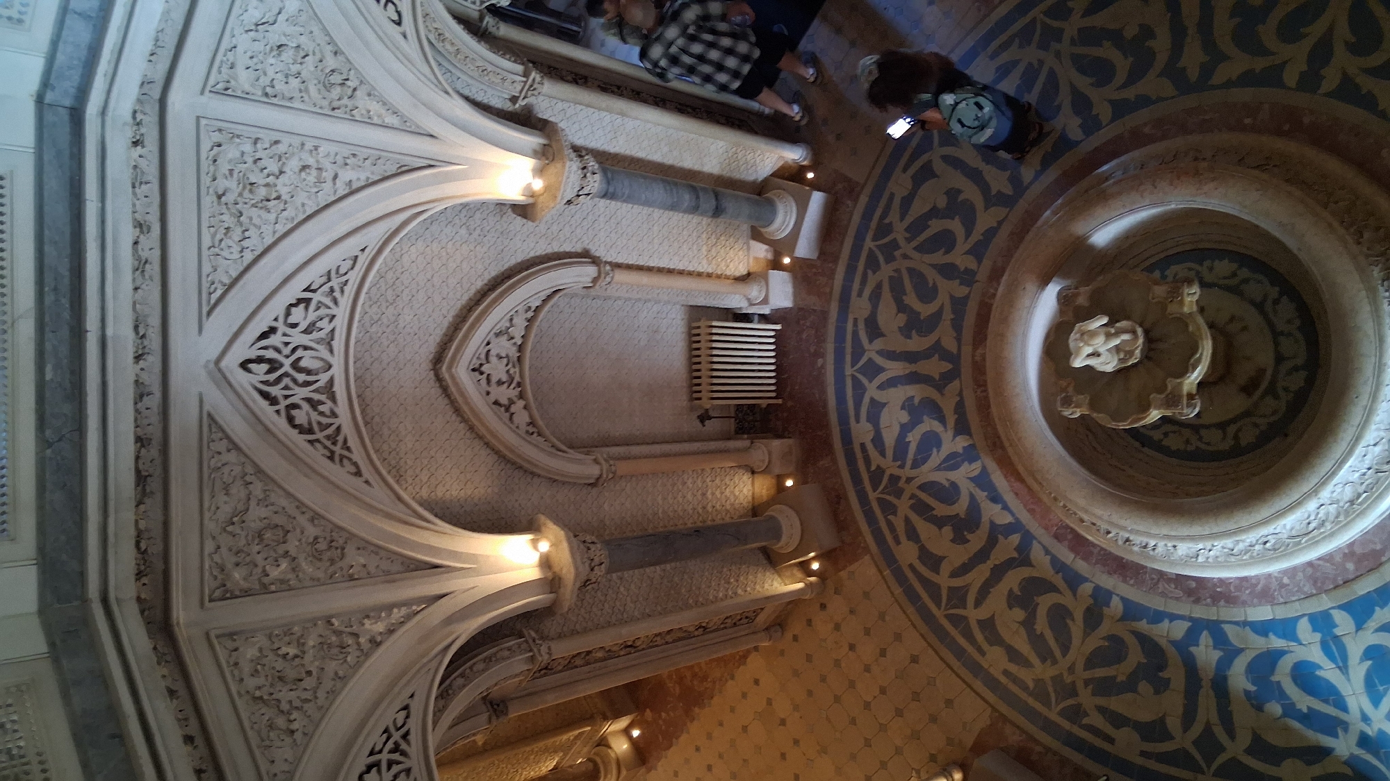This is a high-angle, full-shot image of an interior space with ornate architectural features and a decorative tiled floor. In the center of the room, a circular fountain with a small sculpture is visible. The floor is covered in a geometric mosaic pattern with blue and tan tiles, bordered by a red and blue band. Above the fountain, intricate white carved stucco work adorns the ceiling and arches. Two large, fluted columns with grey marble shafts and ornate capitals support the arches. A white radiator is mounted on the wall of an alcove.
On the right side of the image, a person wearing a plaid shirt and shorts is walking away from the camera, holding a phone. Further in the background, another person is visible, also walking away from the camera. Another person, wearing a backpack, is standing near the fountain, looking down at their phone. The walls are decorated with a textured pattern. The lighting is warm, with spotlights illuminating the architectural details and the fountain. The overall impression is of an opulent, historic interior.