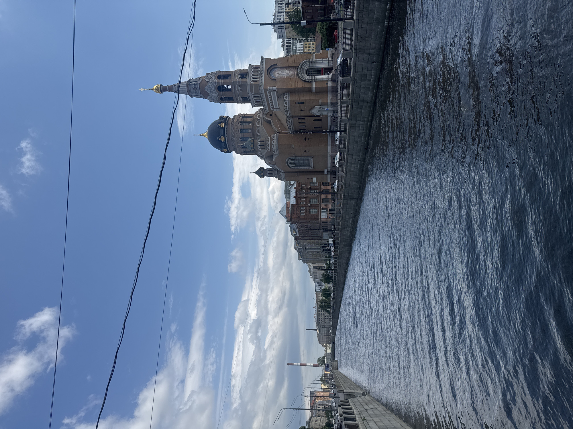 An eye-level, long shot captures a wide canal with dark, rippling water in the foreground. The canal banks are lined with stone retaining walls and metal railings. On the right bank, a large, ornate brick church with multiple domes and a tall bell tower dominates the scene. Cars are parked along the street in front of the church and on the opposite bank. The left bank features a street with buildings, a tall, red and white striped smokestack in the distance, and a bridge crossing the canal further down. The sky above is a vibrant blue with scattered white clouds, and several dark power lines are stretched across the upper portion of the frame. The overall lighting suggests a daytime scene under a partly cloudy sky.