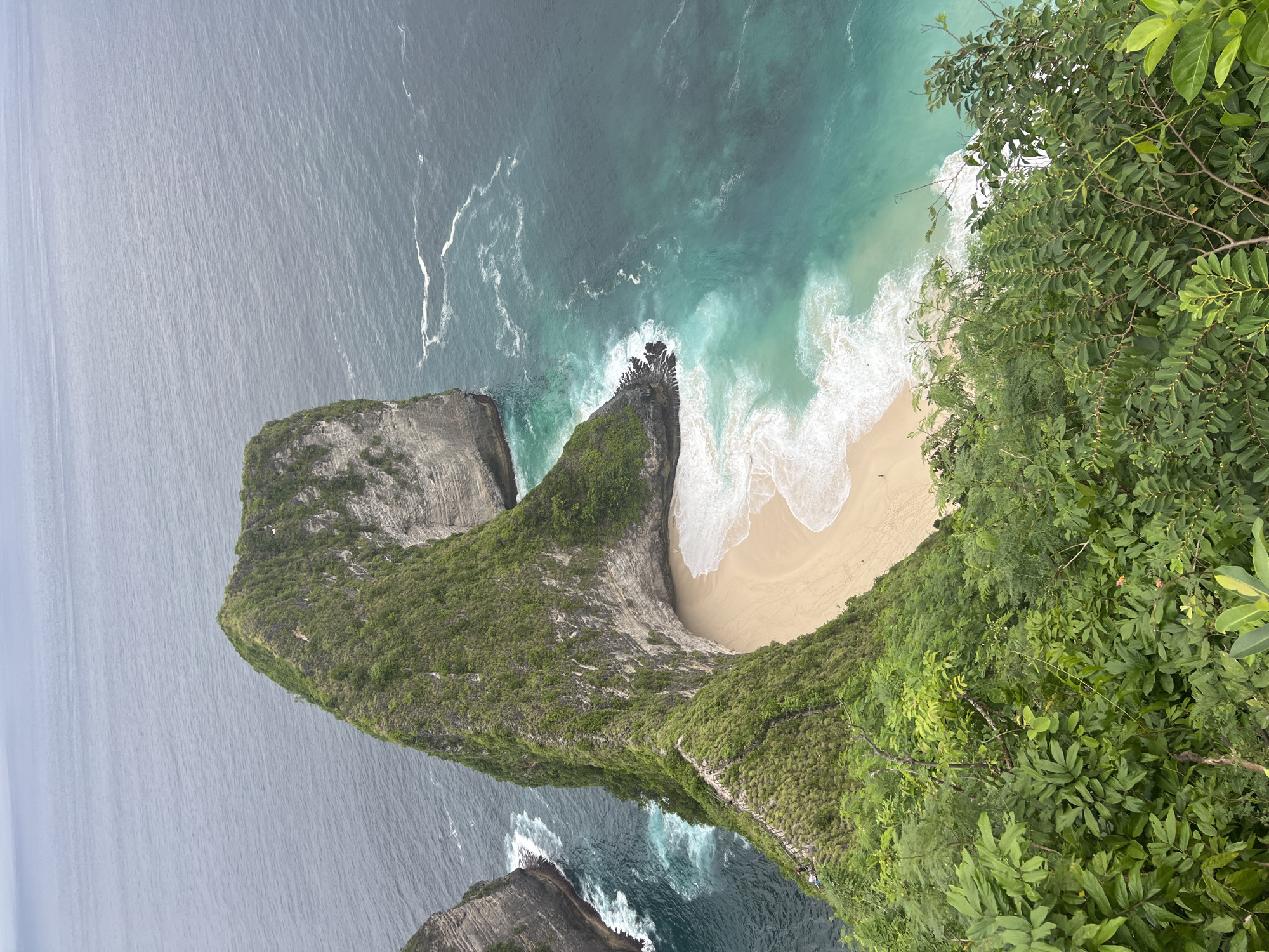 This media file captures a breathtaking aerial or high-angle view of Kelingking Beach in Nusa Penida, Indonesia. The scene is dominated by a dramatic, dinosaur-head-shaped cliff formation, often referred to as T-Rex Bay.
The main subject is this iconic headland, covered in lush, dense green vegetation. On the right side of the frame, more vibrant green foliage from a foreground cliff partially frames the view, suggesting the vantage point is from an elevated lookout. Below the headland, a pristine crescent-shaped sandy beach is nestled, bordered by the clear turquoise waters of the ocean.
The ocean itself presents a beautiful gradient of colors, transitioning from deep blue in the distance to a stunning aqua and turquoise closer to the shore, where white foamy waves gently break onto the sand and against rugged rocky outcrops.
Small figures, identifiable as people, are visible on the sandy beach and can also be seen along a winding path carved into the cliffside, leading down towards the beach. This indicates human activity, likely hiking and enjoying the secluded beach.
The lighting suggests an overcast or hazy daytime, as there are no strong shadows and the natural colors are softly diffused. The overall impression is one of majestic natural beauty and a popular tourist destination. No visible text is present in the image.
