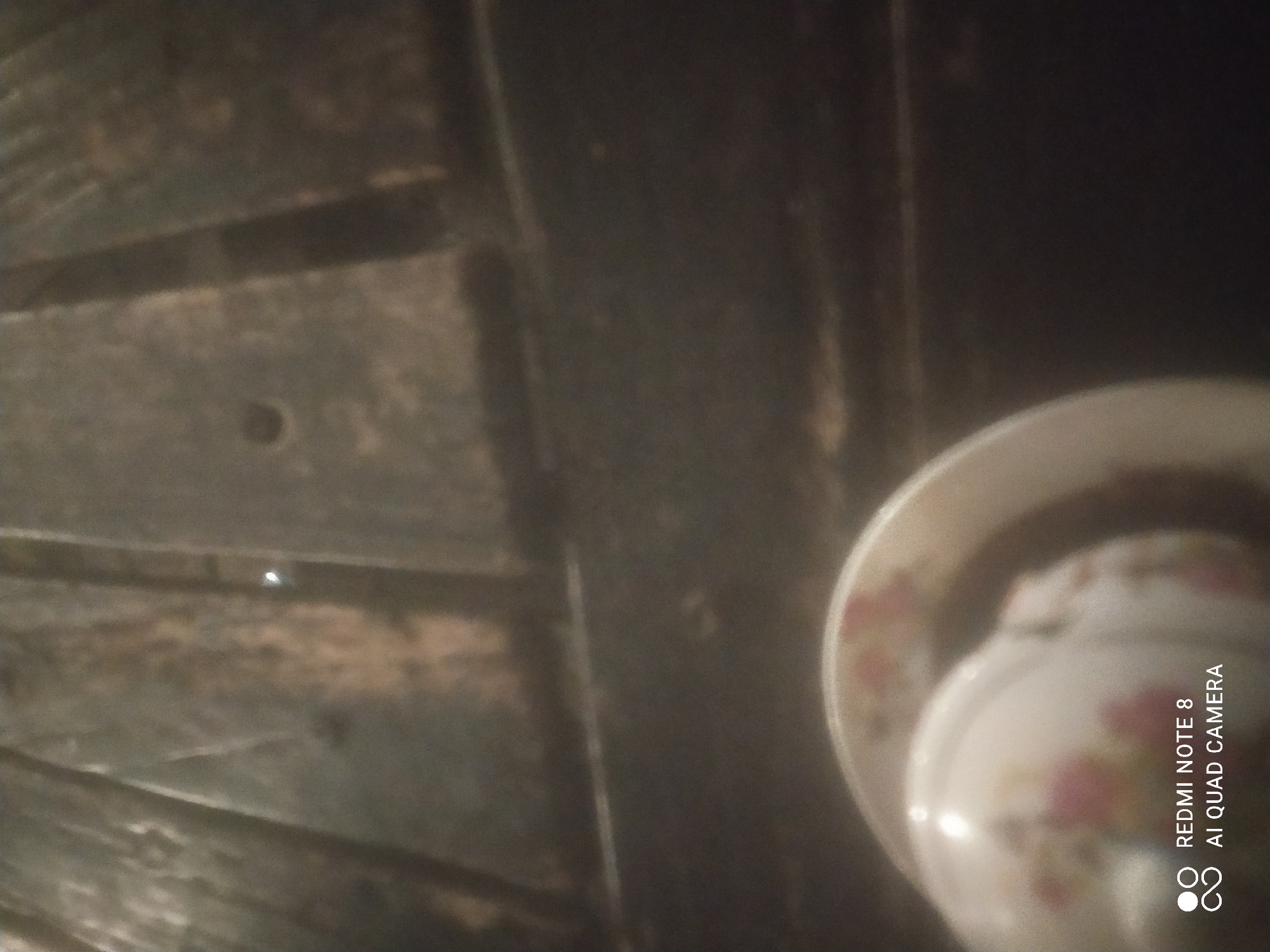 A close-up shot shows a white ceramic cup with a floral pattern. The cup is partially visible in the lower left corner of the frame. The background appears to be a wooden surface with vertical planks. The lighting is dim, and the image is slightly blurred, suggesting it was taken indoors in low light conditions. The bottom left corner of the image contains text overlay which reads "REDMI NOTE 8 AI QUAD CAMERA". There is no identifiable location in the city of Sumurber, Indonesia.