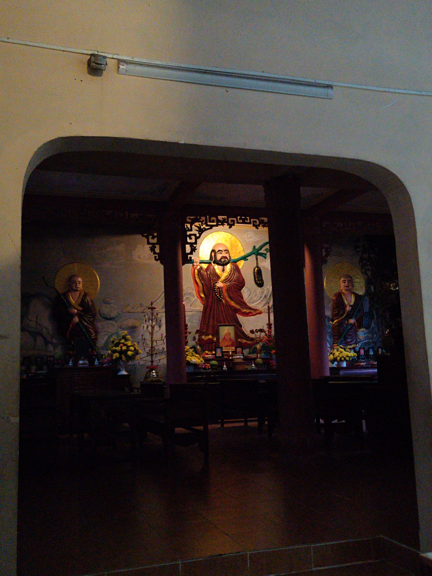 The image portrays an interior view of a temple or pagoda, likely located in Lâm Viên - Đà Lạt, Vietnam, as seen through a dark, arched entryway. The scene is dominated by three large statues of Buddhist figures or monks, positioned behind elaborate altars.
The central and most brightly illuminated figure is a bearded statue wearing a flowing reddish-brown robe and a prominent golden halo. He is depicted holding a staff over his left shoulder, from which a green gourd-like object is suspended. This statue stands in front of a painted backdrop featuring stylized elements, possibly mountains and celestial bodies. Above the central figure's head, some vertical, stylized Asian script is visible. The altar in front of this statue is adorned with offerings, including arrangements of yellow flowers, candles, and a small framed picture or portrait.
To the left of the central figure, another statue with a golden halo, dressed in a darker robe, holds a staff and stands against a mural of natural elements like rocks or clouds. Its altar also holds yellow floral arrangements and a unique, small tree-like decorative item. A third similar statue, also with a golden halo and dark robe, is positioned to the right, likewise holding a staff and flanked by an altar with yellow flowers.
The lighting within the space is dim overall, with strategic spotlights highlighting the statues and their halos. Overhead, simple fluorescent light fixtures are attached to the ceiling, one of which appears to be missing a cover or damaged. Dark wooden pillars frame the central display, creating a structured and reverent atmosphere. The foreground and floor are dark, with reddish-brown tiled flooring visible towards the bottom of the frame. The scene suggests a peaceful and spiritual environment dedicated to worship or contemplation.