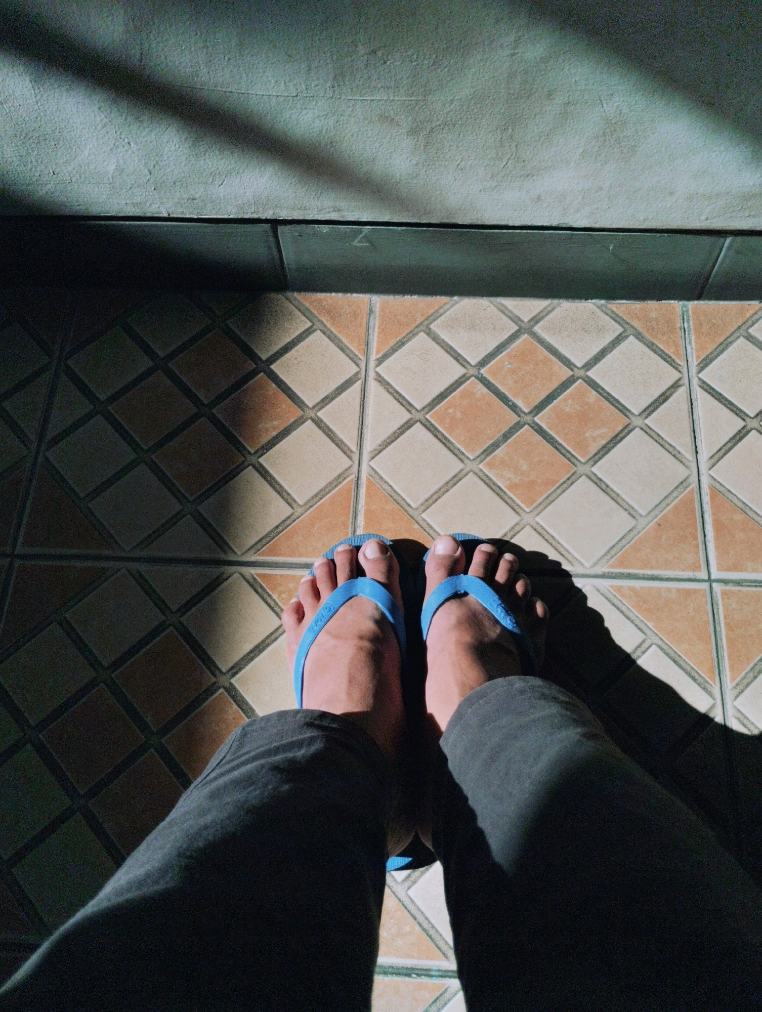 The image shows a pair of feet wearing blue flip-flops, standing on a tiled floor. The tiles are square with a diamond pattern of brown and cream colors. A shadow from a diagonal object falls across the wall and floor. The scene is lit by sunlight. The image does not provide any specific details to determine a location in Ponorogo, Indonesia.