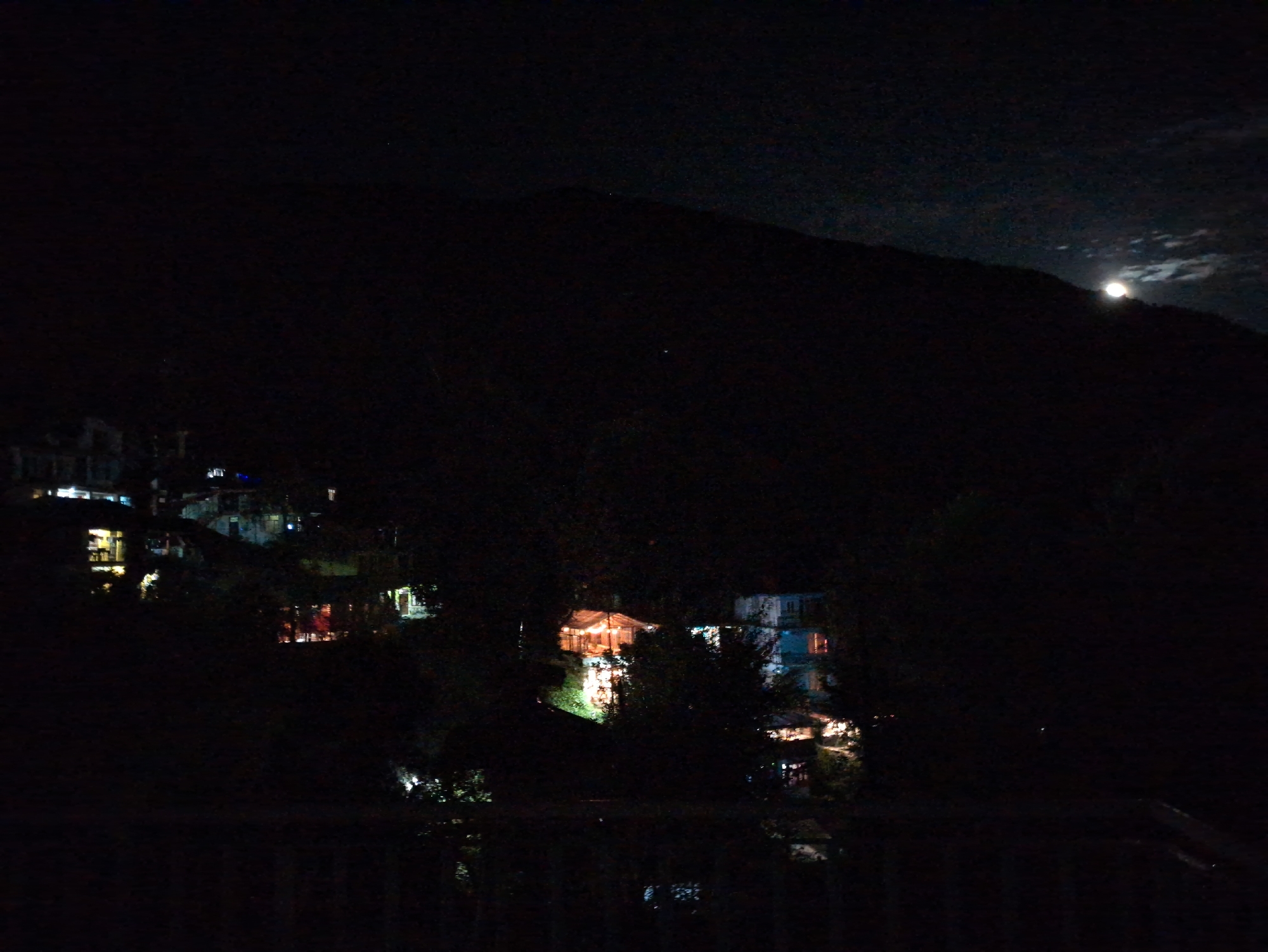 The image depicts a nighttime scene in Bhagsu, India, characterized by darkness and the glow of artificial lights. A large, dark mountain or hillside dominates the background, with the moon visible in the upper right quadrant, partially obscured by clouds. Several buildings are scattered across the lower portion of the hillside, their windows and exteriors illuminated by warm, yellow and white lights, suggesting they are residences or establishments. These lights create a contrast against the deep black of the night sky and the unlit portions of the landscape. The foreground appears to be a dark railing or balcony, implying the vantage point from which the photograph was taken. The overall atmosphere is serene and quiet, indicative of a tranquil night in a mountainous region.