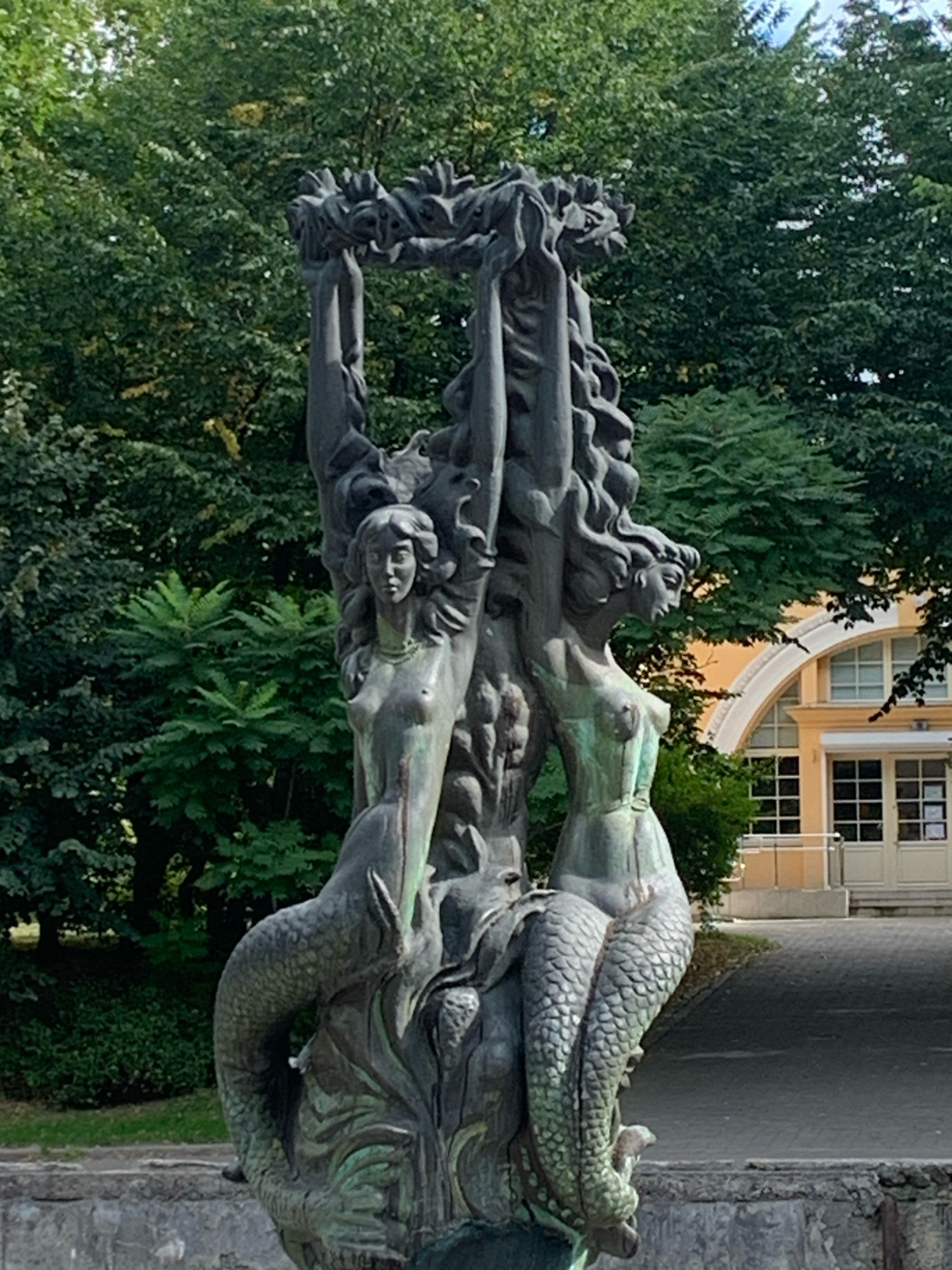 The image shows a bronze sculpture of three mermaids with fish tails, holding a leafy wreath above their heads. The sculpture is located in a park-like setting with mature trees in the background. Behind the sculpture, a building with a light yellow facade and arched windows is visible. The sculpture is weathered, with patches of green patina. The mermaids are depicted with flowing hair and nude torsos. The image is taken from a medium shot, with the sculpture being the central focus.