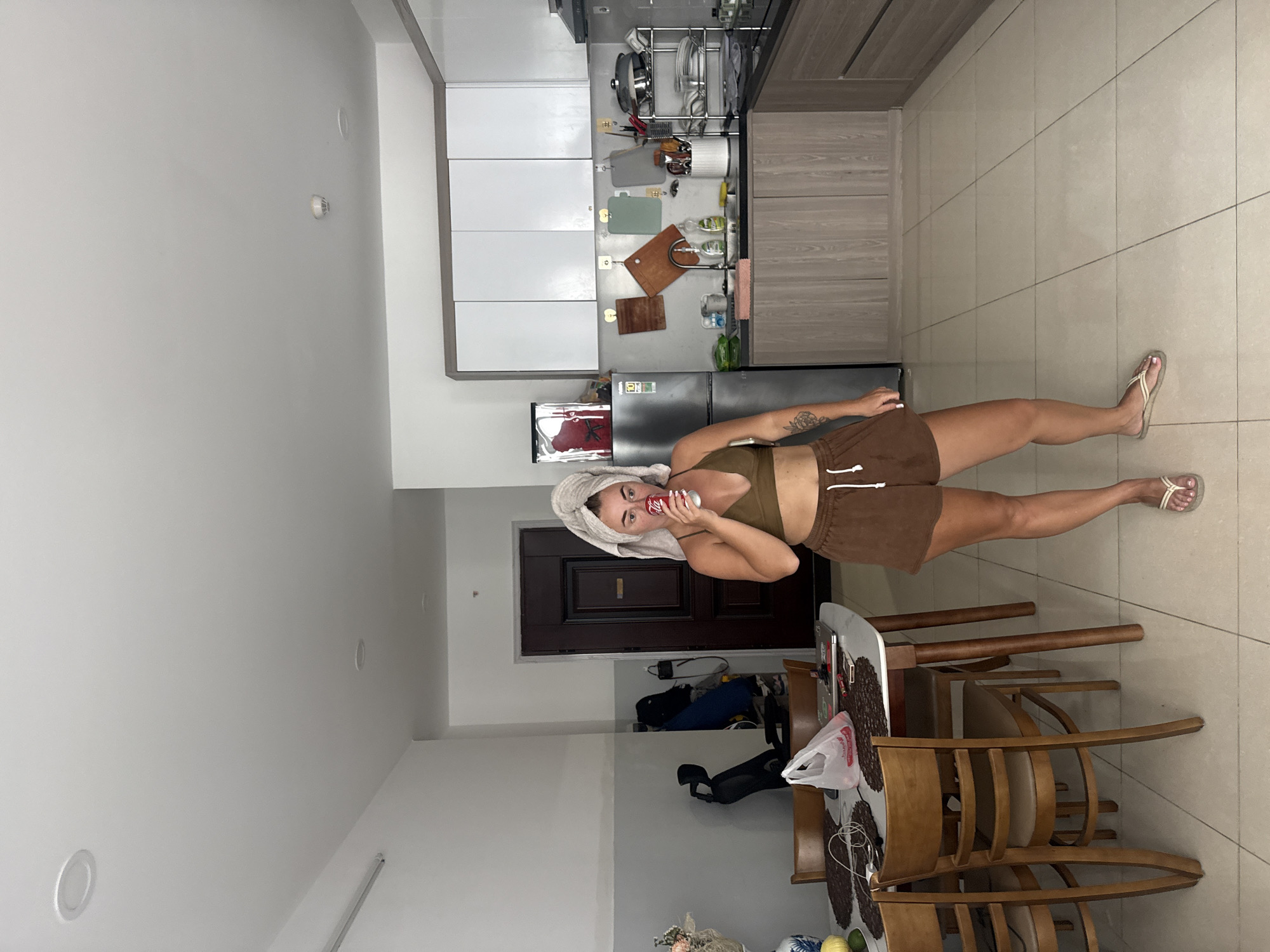 A woman with a towel wrapped around her head is standing in a kitchen, drinking from a can of Coca-Cola. She is wearing a brown sports bra and brown shorts. To her left, there is a dining table with chairs, a plastic bag, and a laptop. The kitchen has wooden cabinets, a refrigerator, and a sink area with various utensils and cutting boards. The floor is tiled. This scene is set in an apartment in Nha Trang, Vietnam.