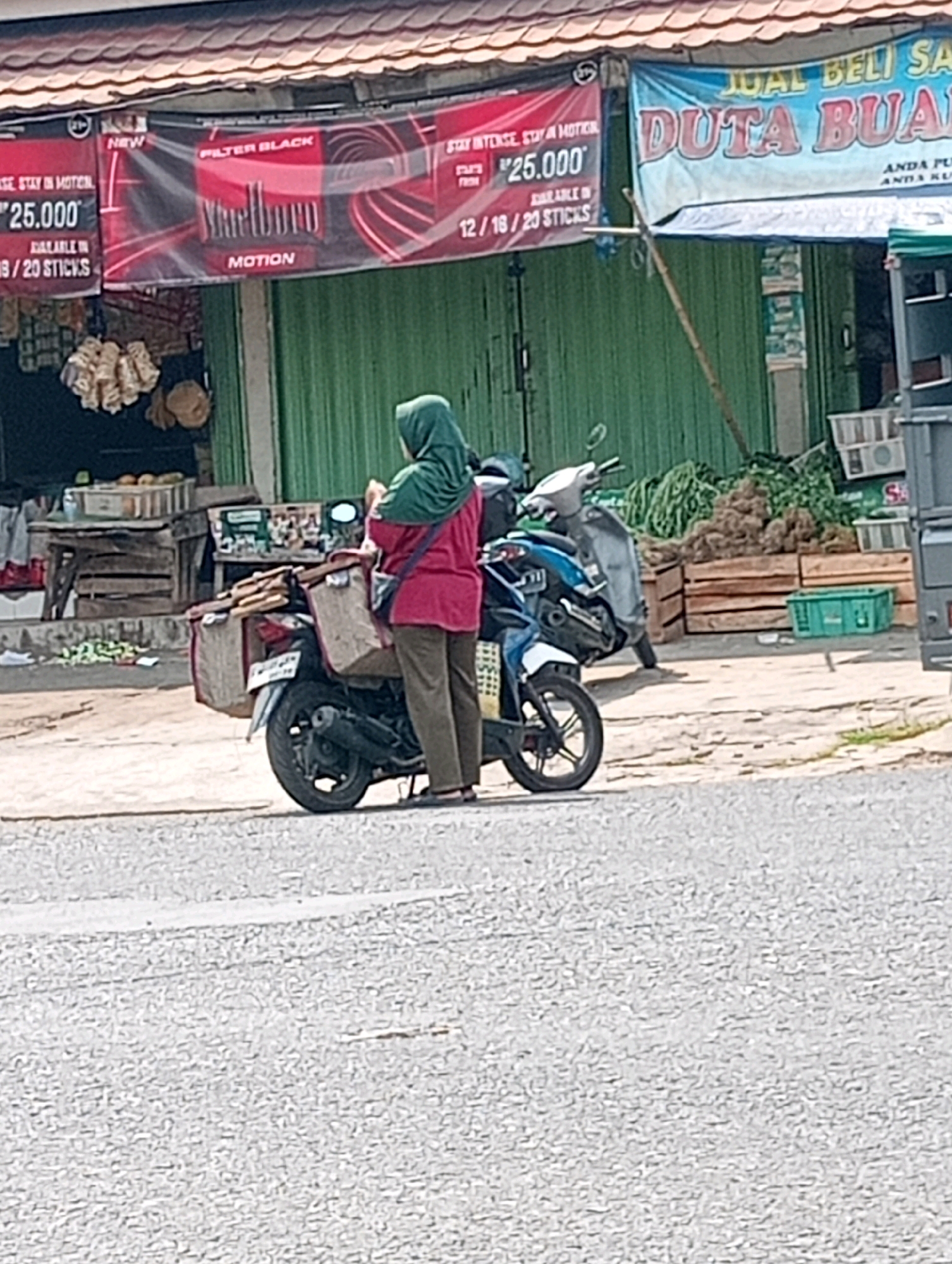 A woman wearing a green hijab and a red shirt stands beside a parked motorcycle in what appears to be a street market. The motorcycle has saddlebags on either side, one of which contains items. In the background, there is a green metal storefront with various advertisements and signs. One sign, featuring a Marlboro logo, promotes cigarettes. Another sign, in blue and white, reads "JUAL BELI SA DUTA BUAH" which translates to "SALE BUY FRUIT AMBASSADOR". Wooden crates filled with produce are visible behind the motorcycle, and some items are hanging on the left side of the storefront. The ground is paved with asphalt.