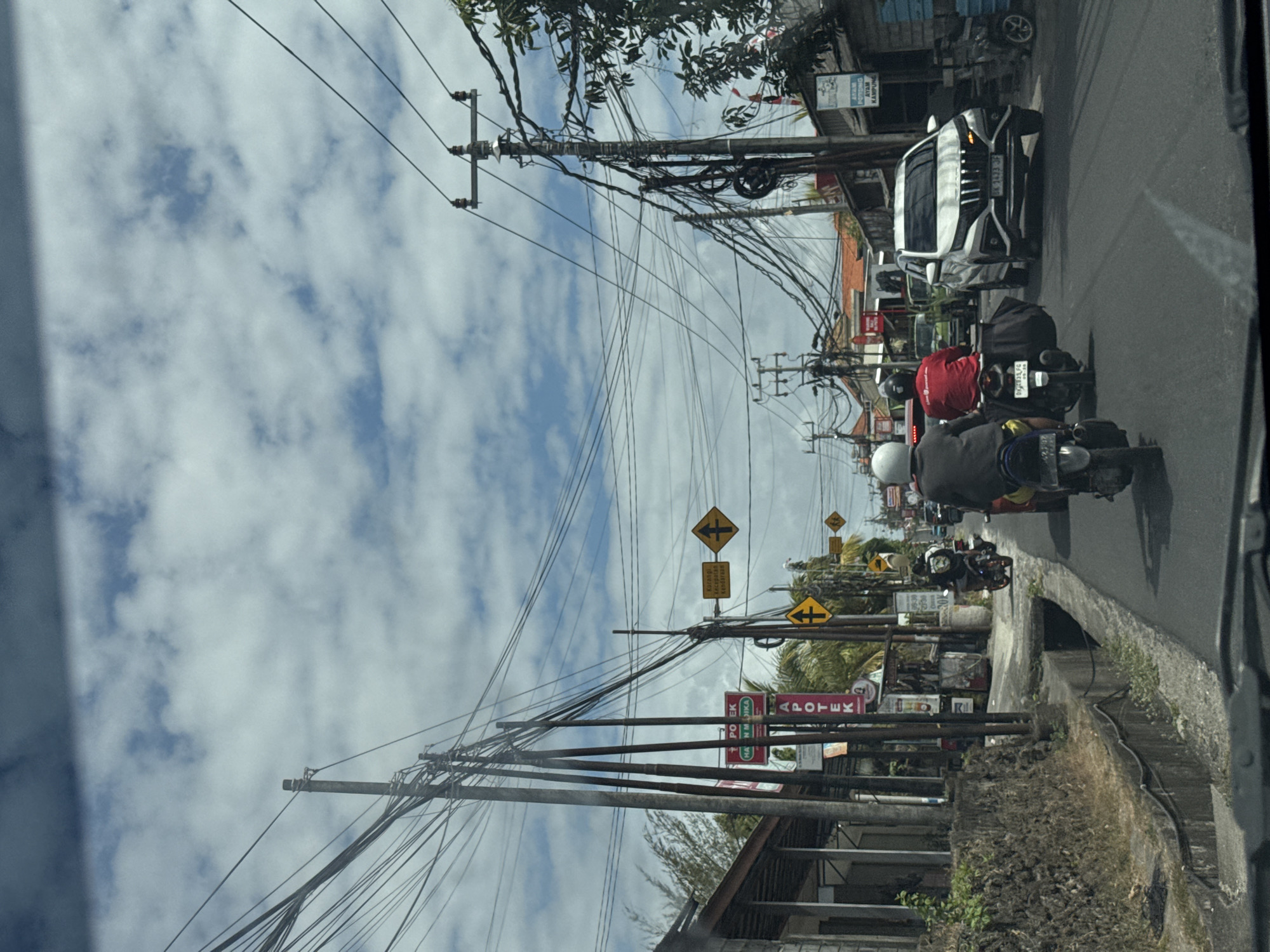 A view from a vehicle traveling on a paved road in Kuta Selatan, Indonesia, shows a cloudy sky overhead. The road is lined with buildings and electrical poles with numerous wires. Two individuals on motorcycles are in the foreground, with one white SUV and a green truck visible further ahead. Yellow traffic signs are posted along the roadside. The overall scene depicts a typical street in a developed urban area.