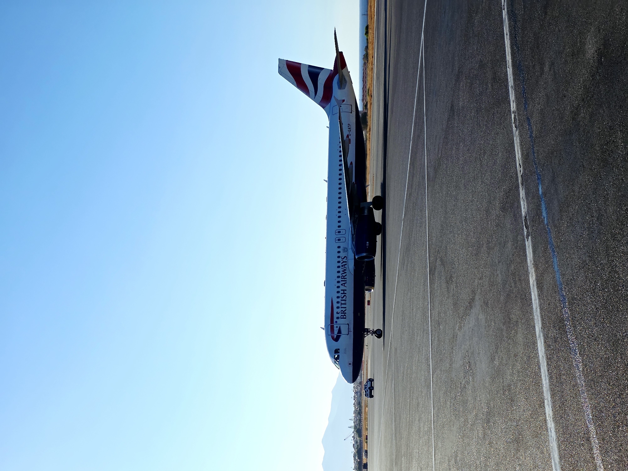 An Airbus A320 aircraft operated by British Airways, with registration G-GATP, is positioned on a tarmac at an airport. The aircraft is white with blue and red livery, featuring the "BRITISH AIRWAYS" text on its fuselage and the Union Jack motif on its tail. The plane is facing towards the right of the frame, and its landing gear is extended. A small white vehicle, possibly a ground support vehicle or a car, is visible in the distance to the left of the aircraft. The tarmac is paved and marked with white lines. In the background, beyond the tarmac, there is a strip of dry vegetation, followed by a body of water, and then distant hills or mountains under a clear blue sky. The scene suggests an airport environment in a sunny location, potentially near a coastal area, given the presence of the sea. The image captures the aircraft at what appears to be a stationary moment on the ground, possibly taxiing or waiting. The location is identified as Nea Alikarnassos Municipal Unit, Greece.