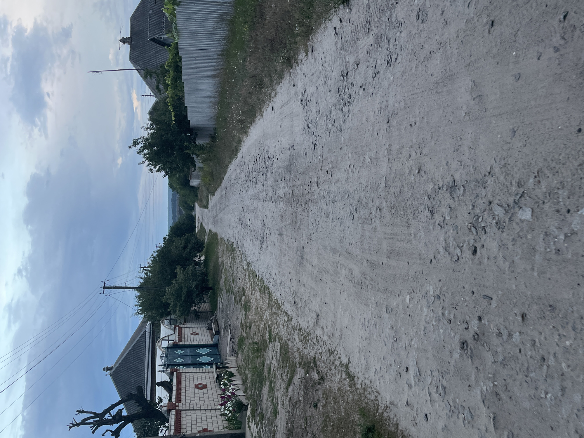 The image depicts a rural dirt road in Vvedenka, Ukraine, under an overcast sky. The road is unpaved and shows signs of wear with loose stones and gravel. On the left side of the road, there is a brick wall with a decorative pattern and a green metal gate. Adjacent to the wall are flowerbeds with blooming white and purple flowers. Further back on the left, a house with a grey, gabled roof is partially visible, along with trees and power lines. On the right side of the road, a corrugated metal fence runs alongside the property, with trees and vegetation growing behind it. The perspective suggests the photograph was taken from a low angle, looking down the road into the distance.
