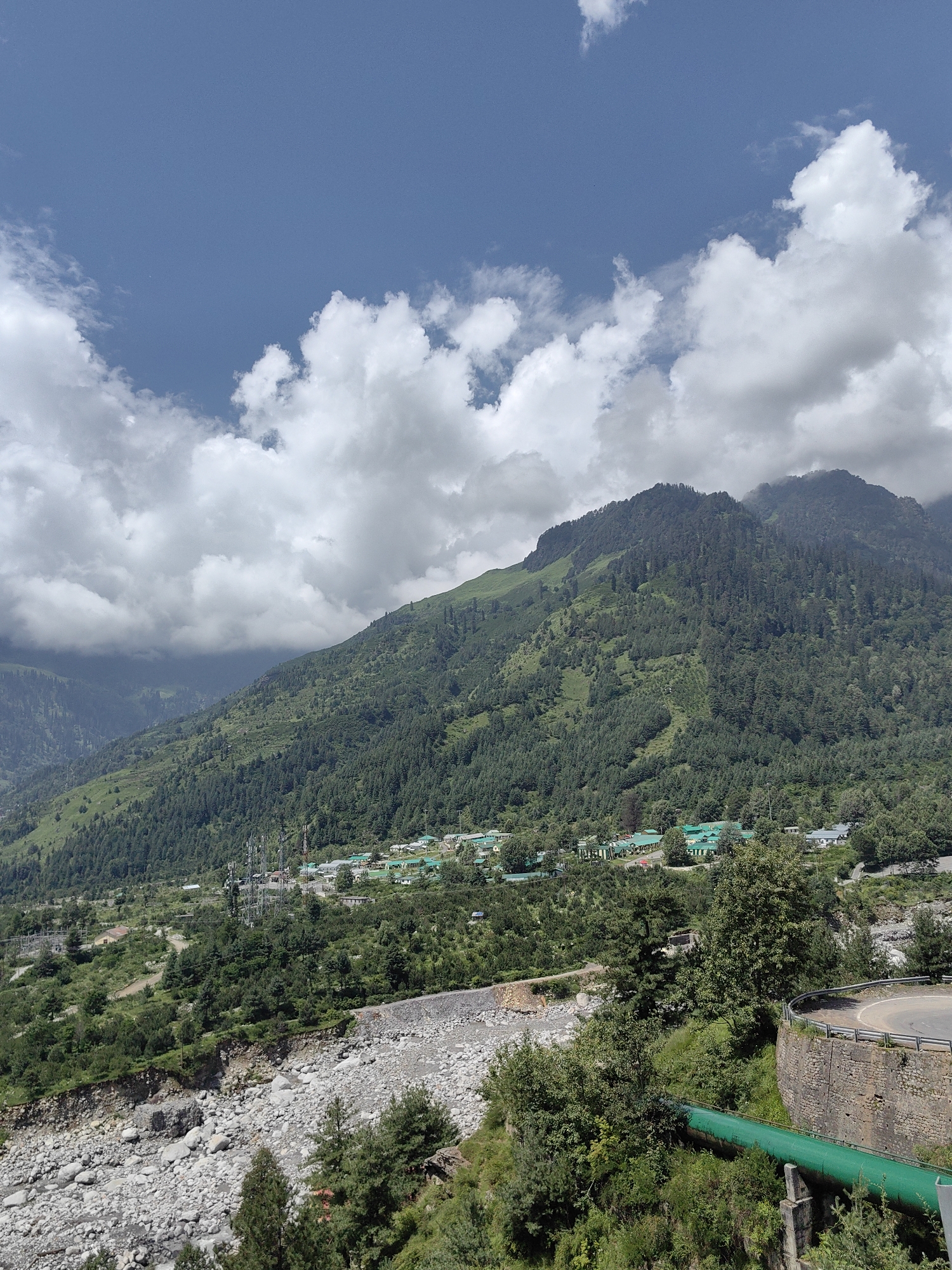 This image depicts a mountainous landscape near Kulang, India, characterized by lush green forests covering the slopes of a large mountain. In the foreground, a rocky riverbed runs along the base of the mountain. A prominent bright green pipe traverses the lower portion of the scene, supported by concrete structures. A paved road curves upwards along the right side, bordered by a stone wall. In the middle ground, a cluster of buildings with green roofs, likely part of a settlement or resort, is visible nestled at the foot of the mountain. Several tall communication towers are also present in this area. The sky is a vibrant blue, dotted with white, puffy clouds. The overall impression is one of a scenic, natural environment with signs of human infrastructure.