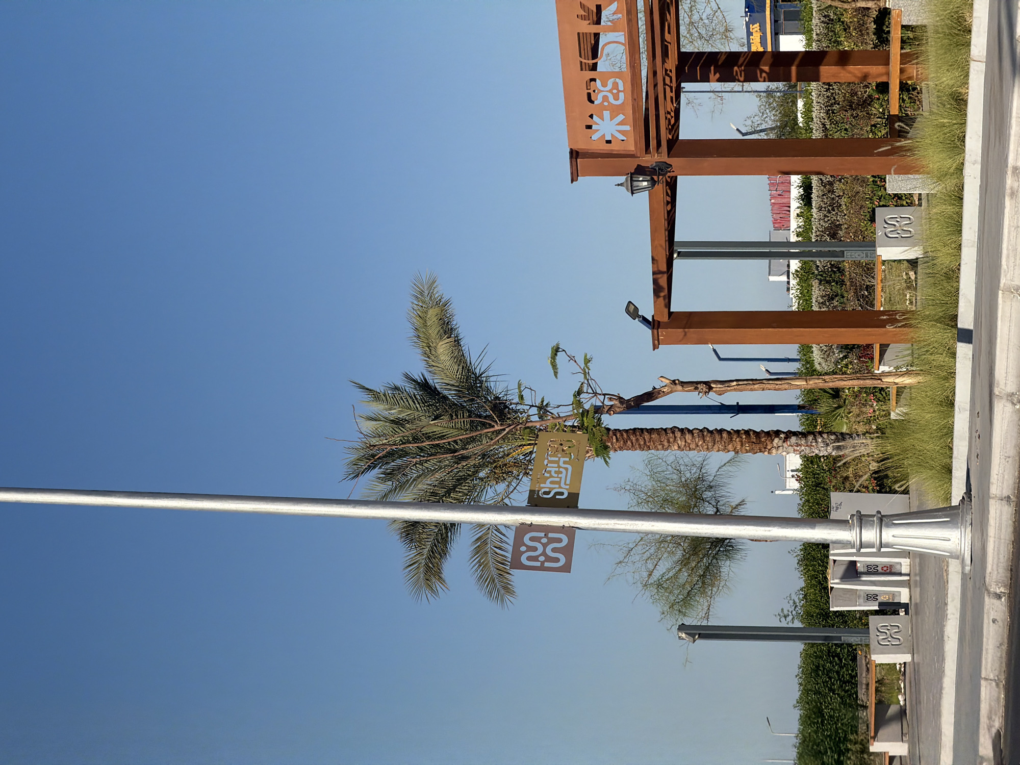 The image depicts an outdoor scene in Sharm El Sheikh, Egypt, on a clear day with a bright blue sky. A tall, silver lamppost stands prominently in the foreground, slightly to the left of center. To its right, a palm tree with lush green fronds is visible, and next to it, a smaller, bare tree trunk. A brown sign with Arabic script and geometric patterns is attached to the lamppost, with a smaller sign featuring a stylized "M" logo below it.
In the middle ground, a rust-colored wooden pergola with intricate geometric cutouts spans across the scene. Two black lanterns are attached to the pergola's structure. Beneath the pergola, benches are placed, suggesting a seating area. To the left of the lamppost, a series of white trash receptacles are arranged. Further back, a dense hedge forms a backdrop, with a few buildings and distant structures visible beyond it. The ground is paved with concrete and bordered by a curb. Lush green grass and ornamental plants are scattered throughout the scene. The overall impression is of a well-maintained public space, possibly a park or a promenade.