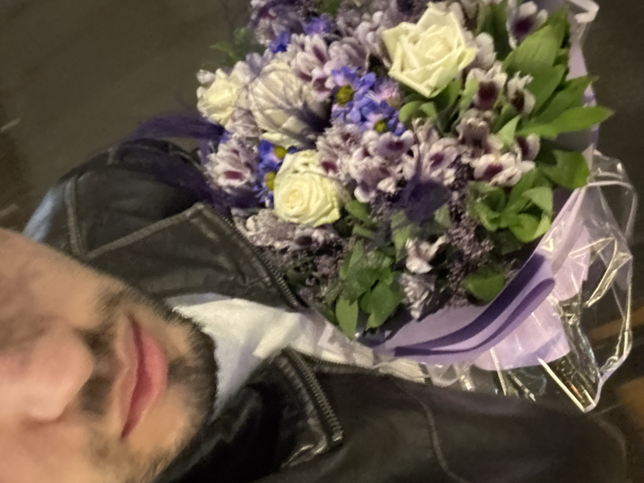 The image presents a close-up of a male subject in Yerevan, Armenia, holding a large floral bouquet. The subject's facial features from the nose to the chin are visible, including a dark beard. He wears a dark leather jacket over a white shirt. The bouquet, occupying the lower right, features white roses, purple and lavender multi-petaled flowers, smaller blue blossoms, and green foliage. It is wrapped in light purple paper and clear plastic. The background is dark and out of focus, indicative of low-light conditions.