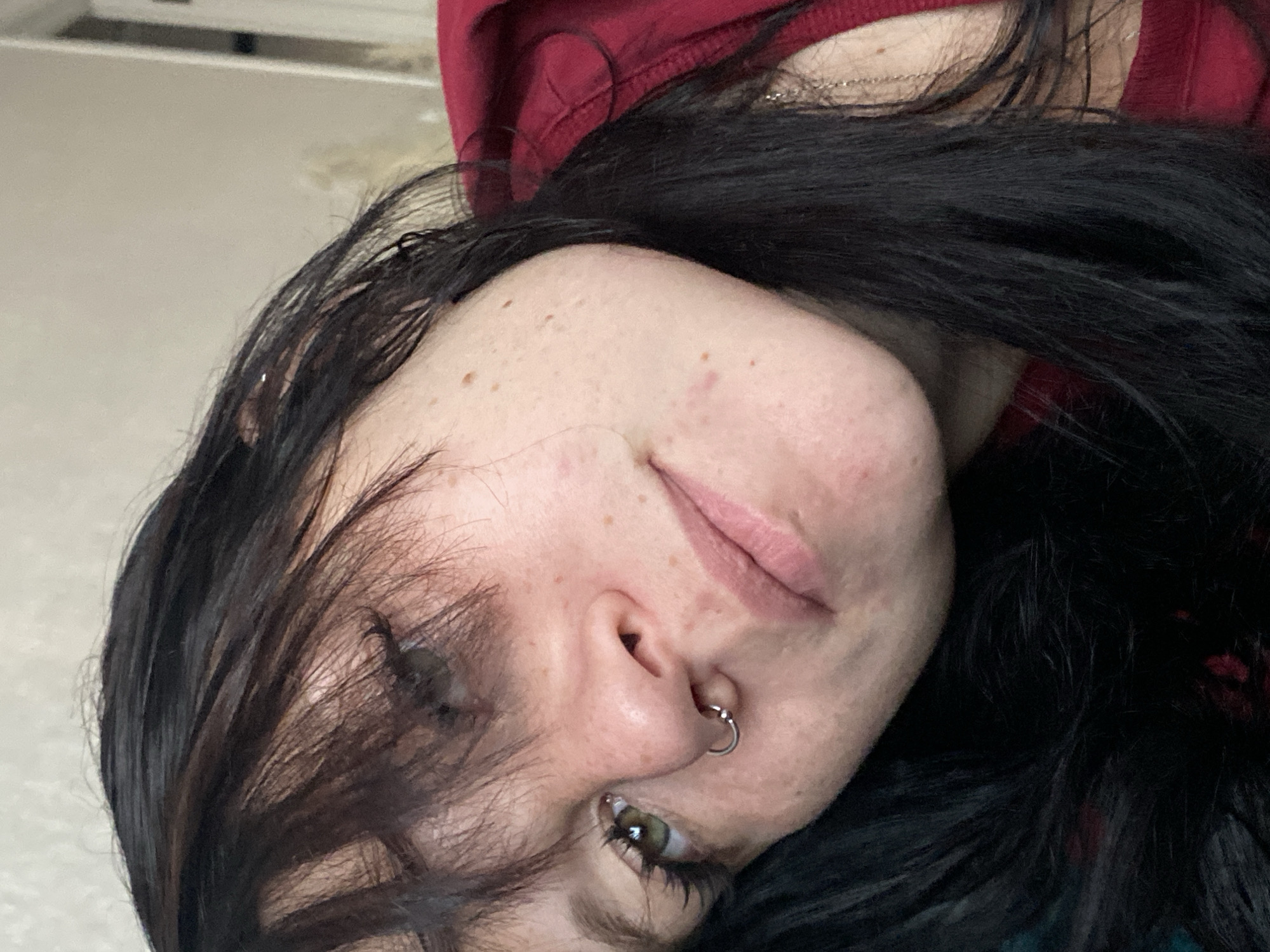 A close-up, overhead shot shows a young woman with black hair and a septum piercing. Her face is tilted to the side, and her eyes are looking off to the left. The woman is wearing a red top. The background is a blurred, neutral wall. No location in Yanino-1, Russia, is visible.