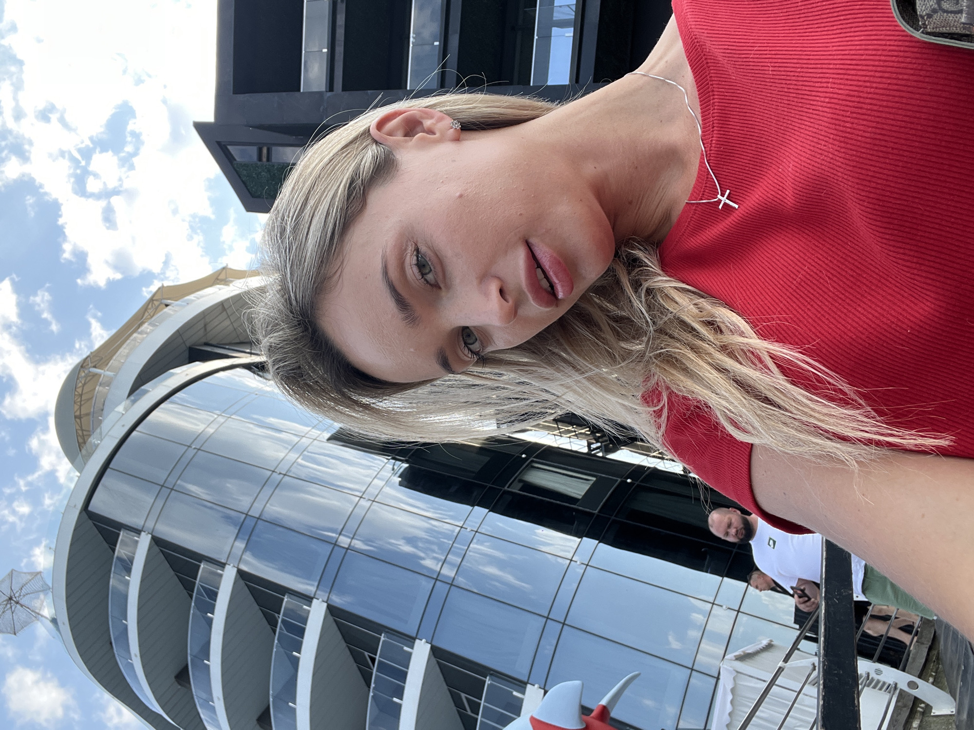 The image shows a woman with blonde hair wearing a red top and a silver cross necklace. She is standing outside in front of a modern building with a glass facade and balconies. The sky is partly cloudy. In the background, there are other people and parts of buildings. This scene is located in Odesa, Ukraine, likely near the waterfront or a modern development area given the architecture.