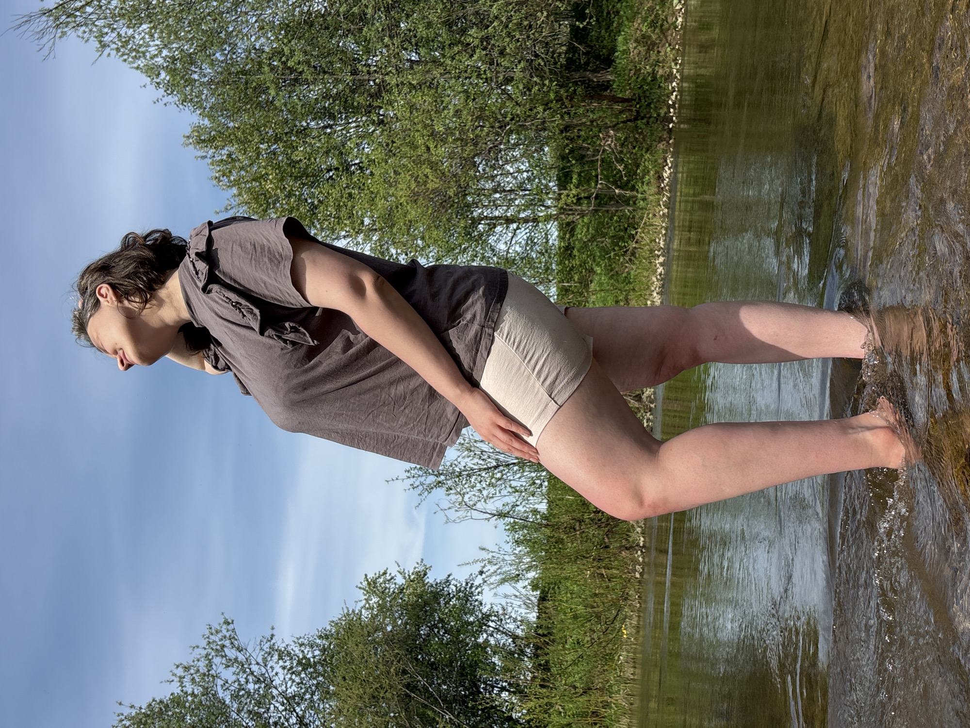 A medium shot shows a woman standing in a shallow body of water, possibly a river or stream, in Усть-Тунтор, Russia. The woman is wearing a short-sleeved, gray-brown shirt and light-colored shorts. She is looking towards the water and is in a slightly bent-over position. The water line reaches around her calves. The background shows green foliage, suggesting a wooded or grassy area. Blue sky is visible above, partly obscured by tree foliage.