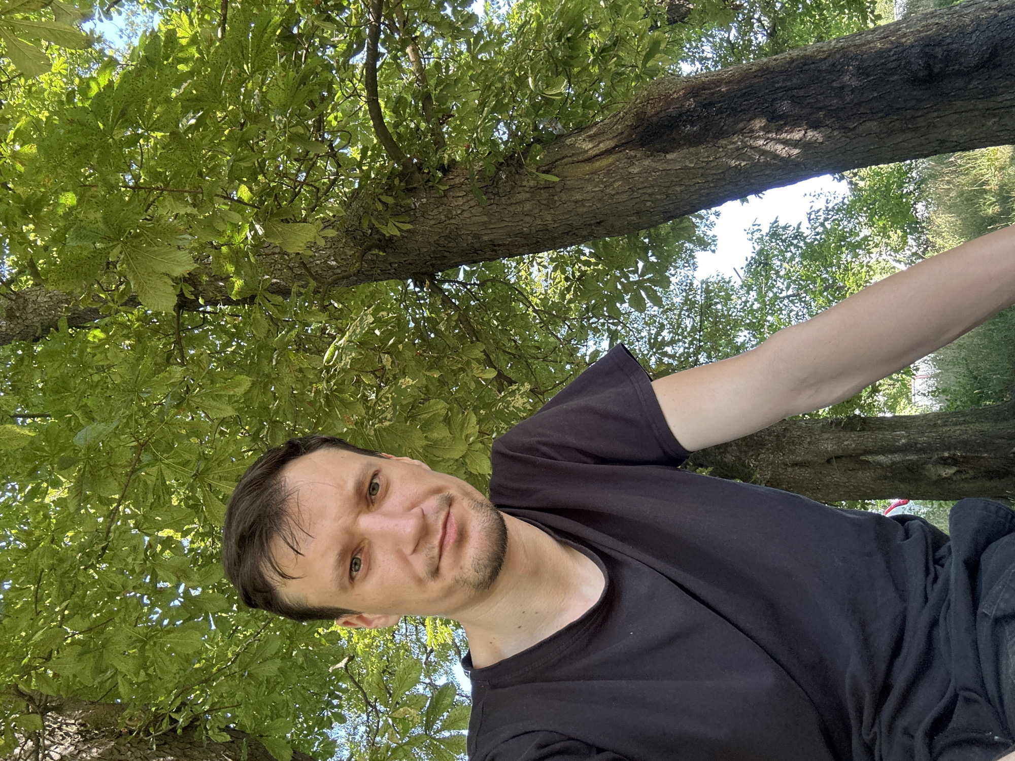 In Makiv, Ukraine, a man is taking a selfie outdoors. He is positioned in the foreground, occupying the left half of the frame. The man has brown hair and a light complexion, with stubble on his chin and jawline. He is wearing a black t-shirt. His right arm extends out of the frame. The background is dominated by lush green trees, their leaves creating a dappled effect with sunlight filtering through. The man is looking directly at the camera with a neutral expression. The lighting suggests it is daytime.