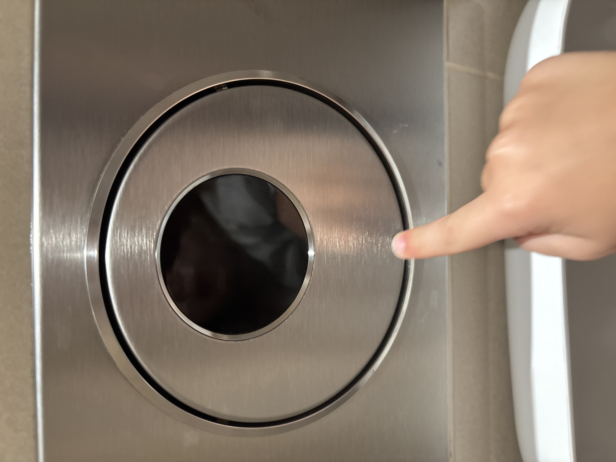 A close-up, eye-level shot focuses on a brushed stainless steel flush plate, likely part of a toilet in a public restroom. The plate features a circular indentation with a dark, reflective interior, possibly a sensor or a decorative element. A child's index finger, with a slightly chipped fingernail, is touching the bottom edge of the plate. The background shows a neutral-toned, tiled wall. The lower portion of the image reveals the edge of a white ceramic toilet bowl. The lighting suggests an indoor, well-lit environment. This scene is located in the city of Dubai, United Arab Emirates.