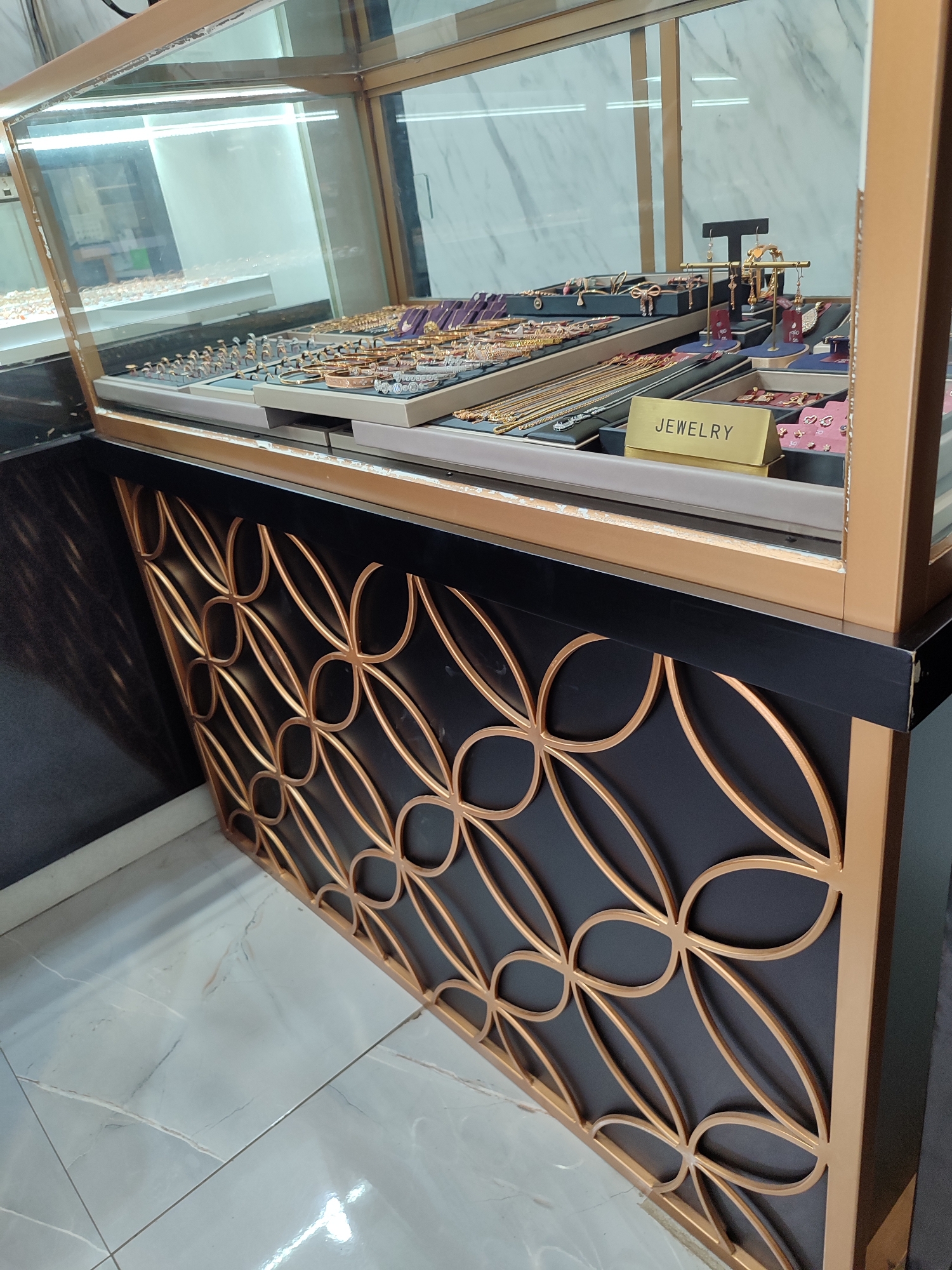 This image depicts a jewelry store display case in Randudongkal, Indonesia. The display case is made of glass and gold-colored metal, and it is filled with various types of jewelry, including necklaces, bracelets, rings, and earrings. The jewelry is arranged on trays and stands, and some of the items are displayed with price tags. A sign with the word "JEWELRY" is visible on one of the trays. The interior of the store appears to be well-lit, with bright lights illuminating the display case. The floor is tiled with a marble-patterned design. The background of the display case shows a marble-patterned wall. The lower part of the image shows the base of the display case, which is made of black material and features a decorative gold metal grill with an ogee pattern. The overall scene suggests a retail environment for fine jewelry.
