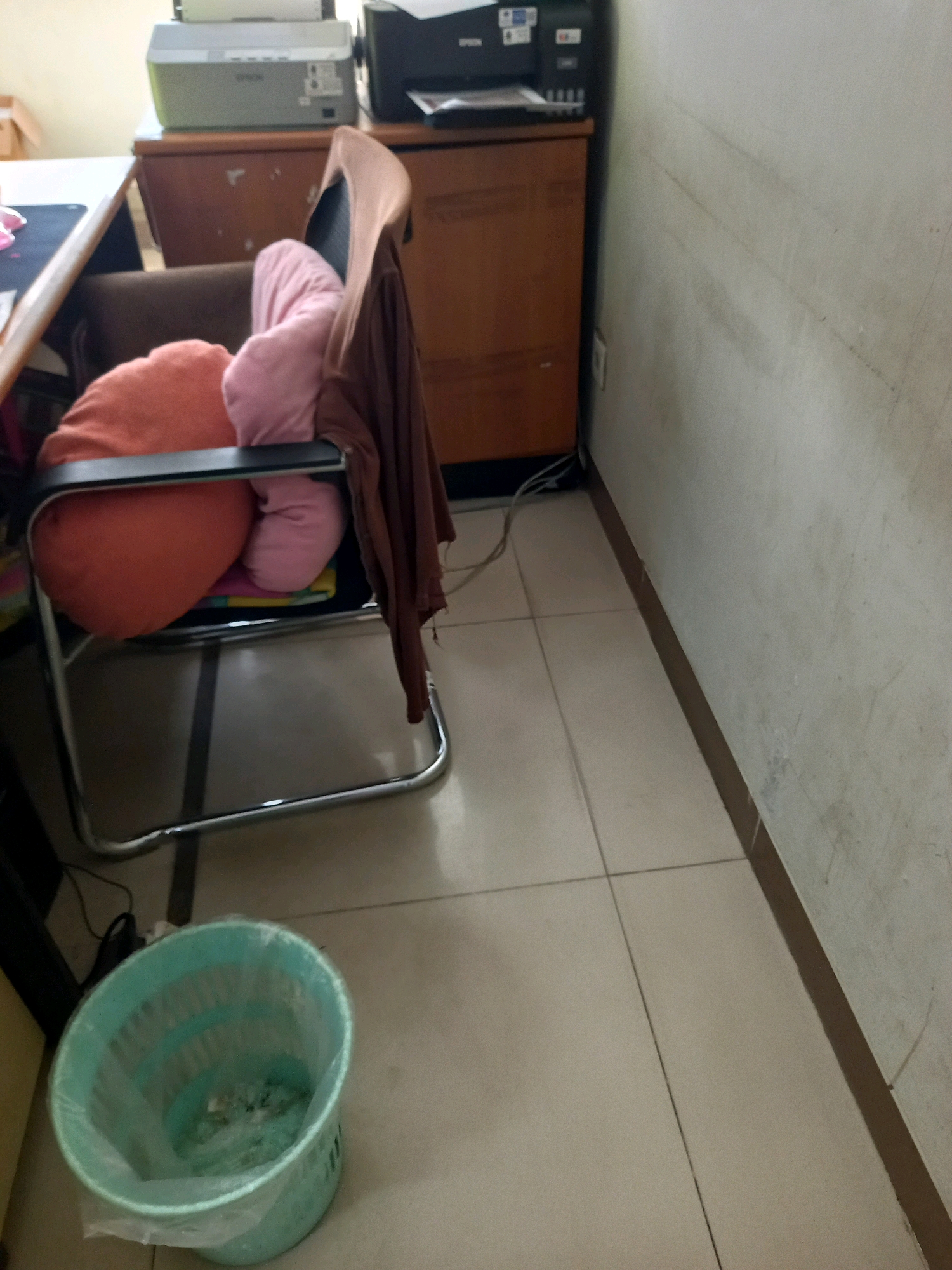 A sparsely furnished office space is depicted in this image, featuring a desk with an Epson printer and an office chair. The chair is covered with two plush cushions, one orange and one pink, and a brown garment is draped over its back. A turquoise trash can with a plastic liner is visible in the foreground, situated on a tiled floor. The scene is likely from an office environment within the city of Samarinda, Indonesia, given the context of the request. The walls are painted a neutral color and show signs of wear, with faint lines and smudges. The lighting suggests it is daytime.