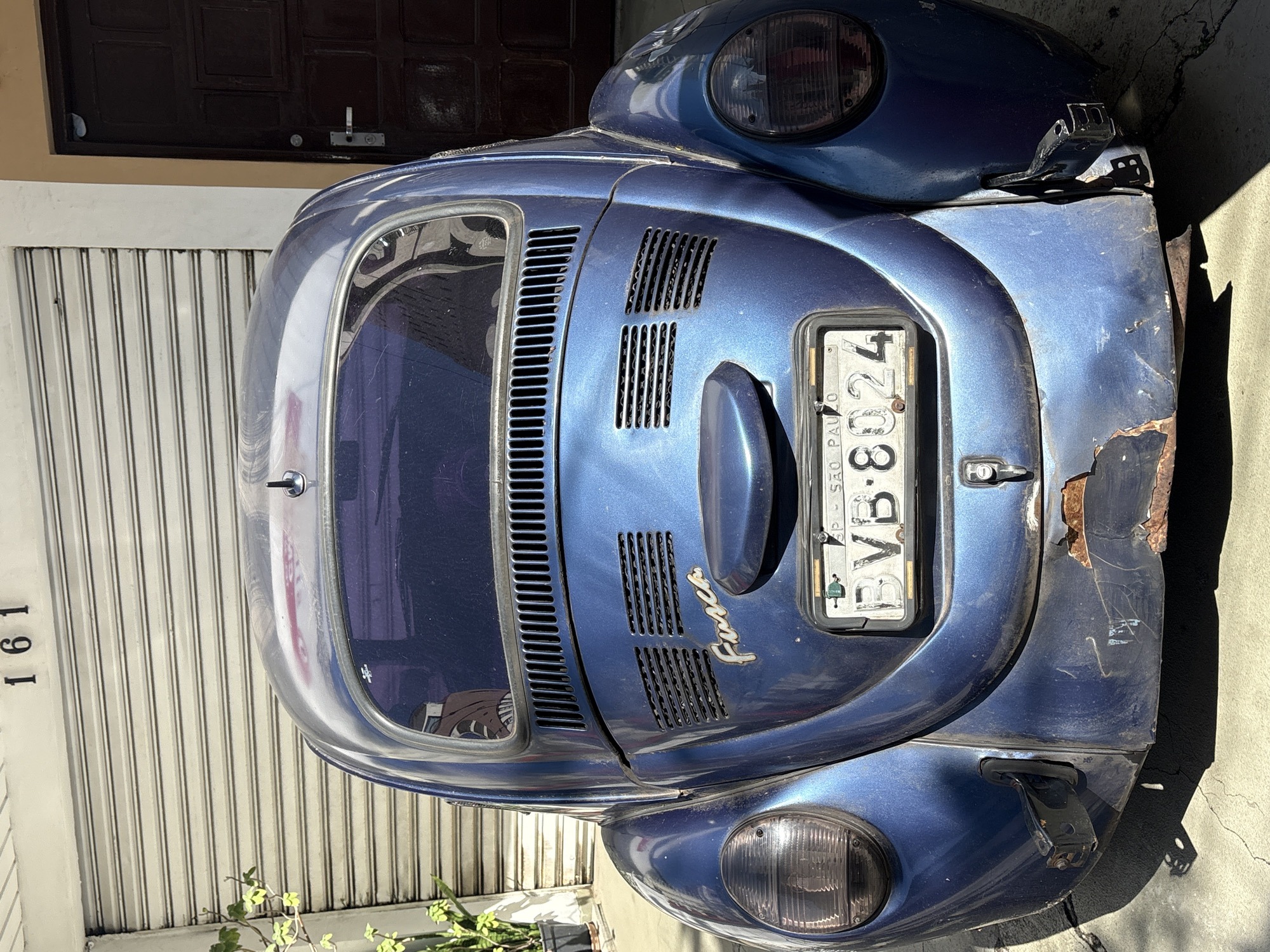 A blue Volkswagen Beetle, known as a "Fusca" in Brazil, is parked outdoors in front of a building. The car's rear is facing the camera, showing its taillights, license plate, and the "Fusca" emblem. The license plate reads "BVB 8024" and indicates "SÃO PAULO". The car shows signs of wear, with rust visible on the lower rear bumper. Behind the car is a corrugated metal garage door, and to the right, a dark brown wooden door. A plant in a black pot is visible to the left of the car. The building's address number "161" is mounted above the garage door. The scene is illuminated by sunlight, casting shadows on the ground. No people are present in the image.