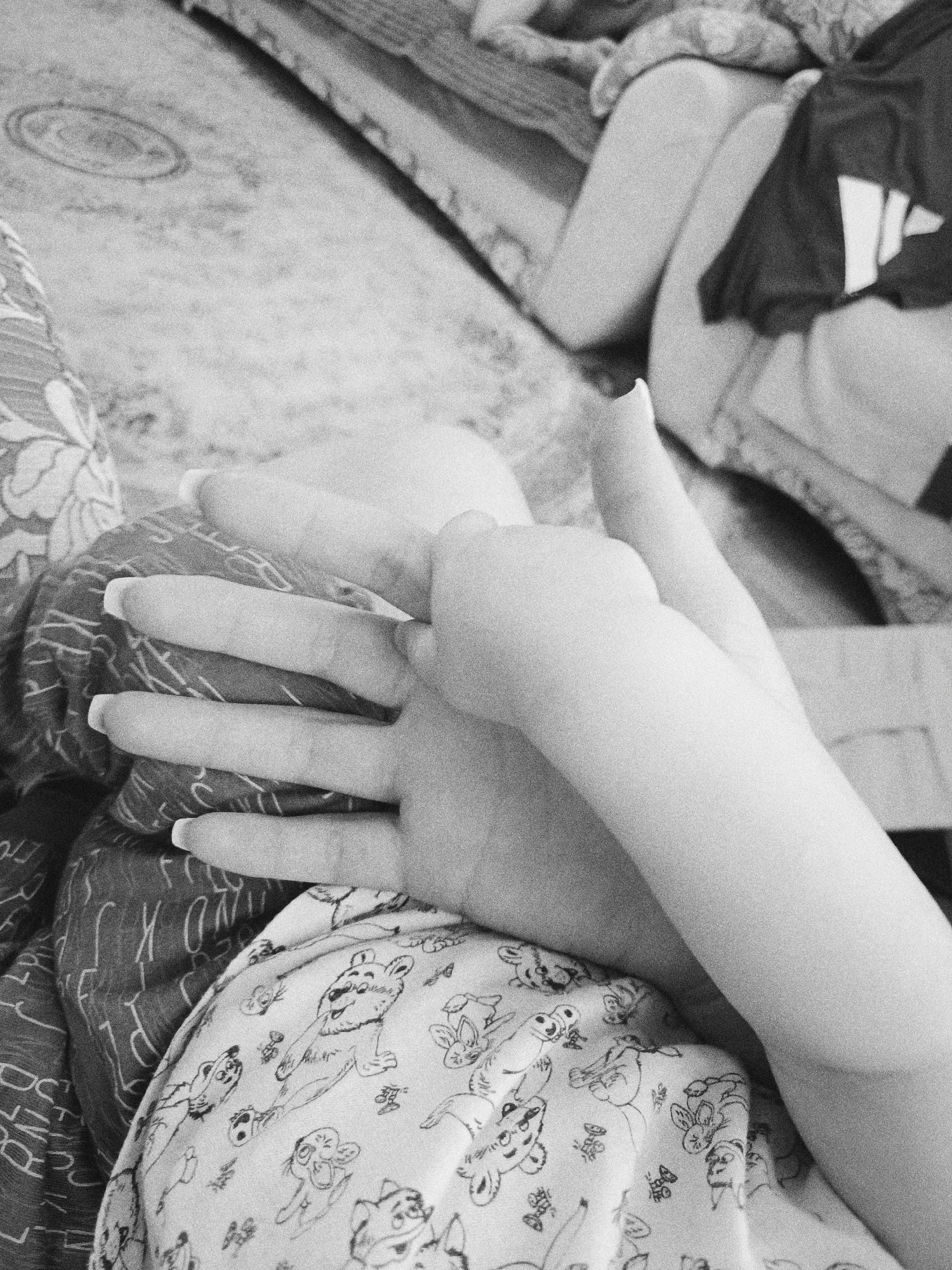 A black and white close-up shows an adult's hand with long nails extending to the left, palm up. A child's hand is clasped within the adult's hand. The hands rest on fabric with cartoon animal patterns. Another patterned fabric is visible underneath. In the upper right corner, a leg is visible, with clothing bunched around it. The location is not specified in the image, nor is it evident from any visual information, therefore I cannot provide any information about it being in Tobolsk, Russia.