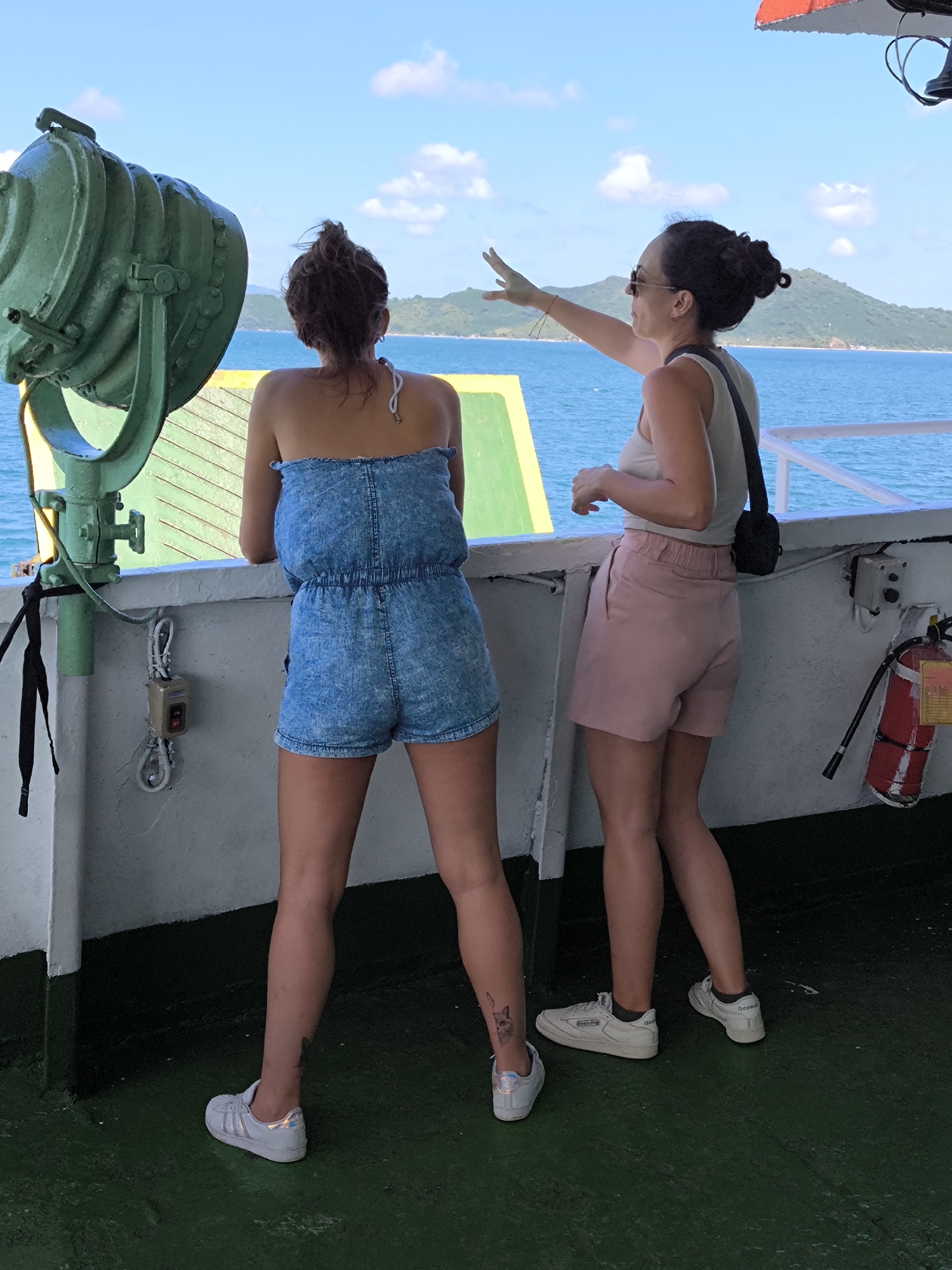 Two women stand on the deck of a boat, facing away from the camera. The woman on the left wears a strapless denim romper and white sneakers. A tattoo is visible on her left leg. The woman on the right wears sunglasses, a light-colored tank top, pink shorts, and white sneakers, with a small black purse hanging from her shoulder. She points her right arm toward the horizon. A large, green spotlight is to the left of the women. A white railing runs along the edge of the boat. Behind the women, there is a view of the ocean, a distant island with mountains, and a cloudy sky. The location is Unknown, Indonesia. A fire extinguisher is mounted on the wall to the right of the women. The deck is green.