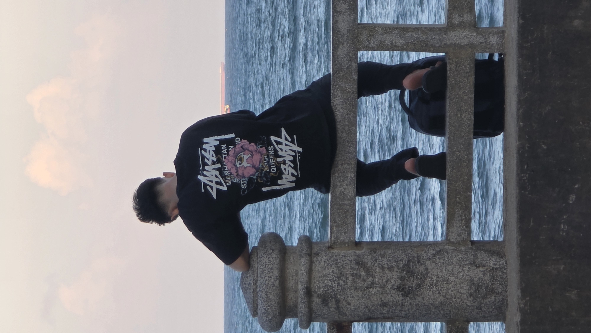 The image depicts a man in a dark-colored Stussy t-shirt, dark pants, and dark socks, standing on a pier or bridge in Nha Trang Ward, Vietnam. He is leaning against a concrete railing, his head bowed. Beyond the railing is a body of water with a horizon visible in the distance. The sky is light-colored with hints of pink and orange, indicating either sunrise or sunset. A dark bag is located on the opposite side of the railing.