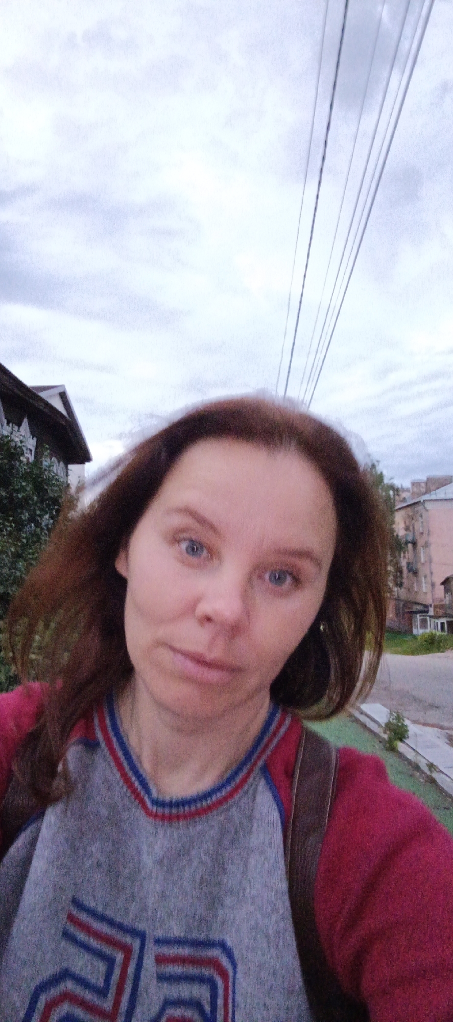 A young adult woman with auburn hair and blue eyes takes a selfie outdoors. She is wearing a grey v-neck sweater with red and blue trim and red sleeves. Behind her, a street is visible with apartment buildings and power lines against a cloudy sky. The scene appears to be in Votkinsk, Russia.