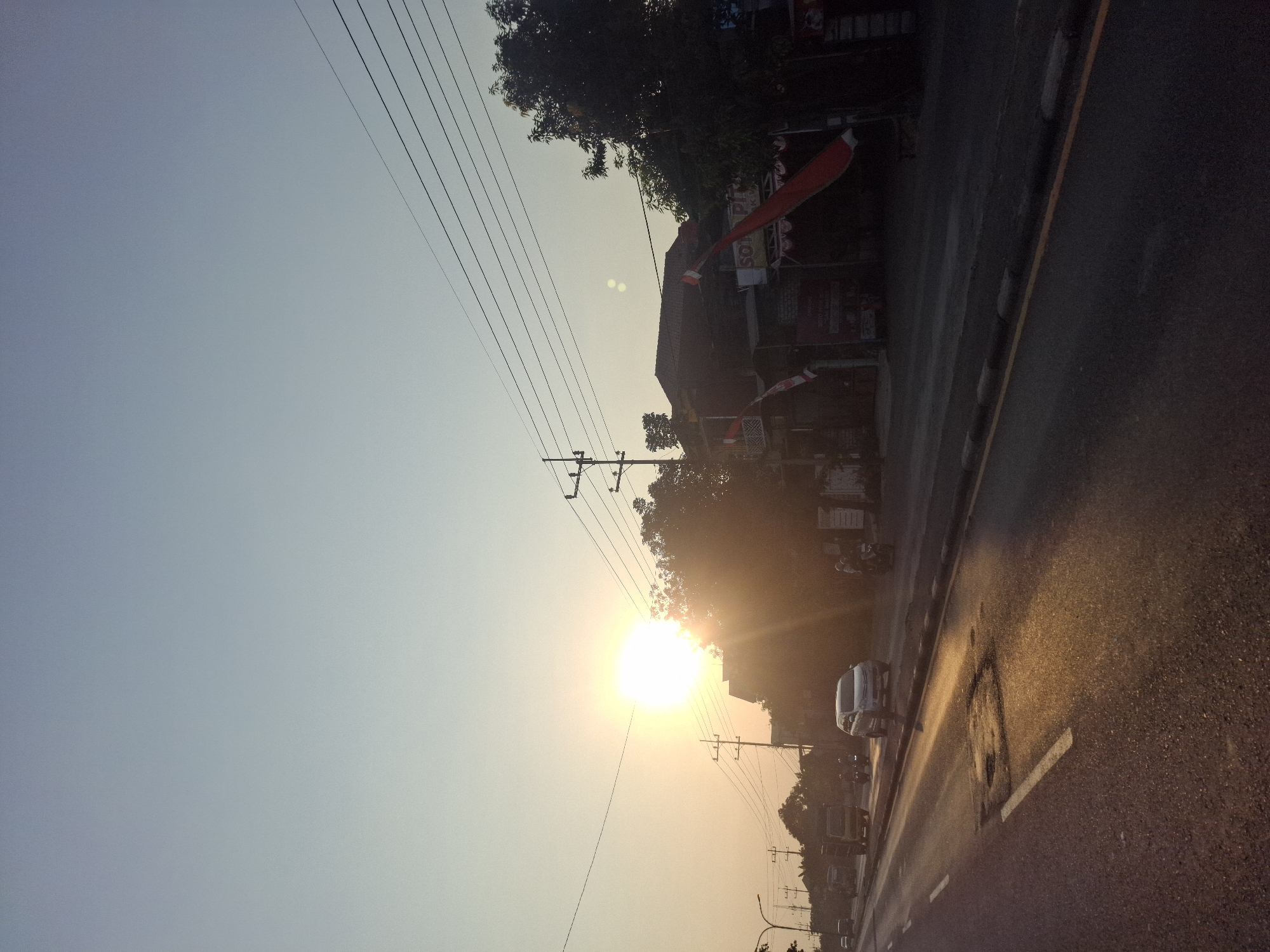The image shows a street scene in Waruwetan, Indonesia, during what appears to be sunset or sunrise. The sun is low in the sky, casting a warm glow and creating lens flares. On the right side of the road, there are buildings with awnings and red and white Indonesian flags displayed. Trees and utility poles with power lines are also visible. Several vehicles, including cars and motorcycles, are on the road. The road surface shows signs of wear, with a visible pothole in the foreground. The sky is a pale blue, fading to a lighter shade near the horizon.