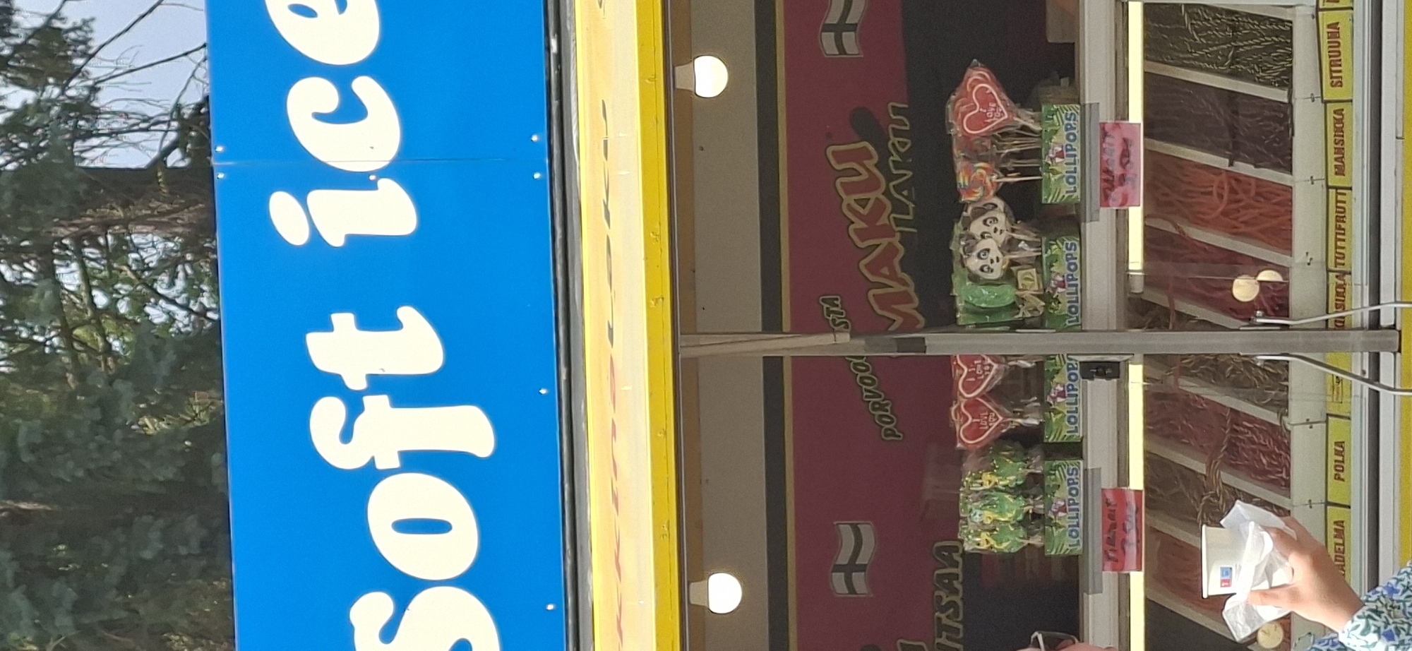 The image shows a street food vendor selling ice cream and candy in the city of Turku, Finland. Above the vendor's stall, a blue sign reads "soft ice" in large, creamy white letters. The vendor's stall is decorated with red banners featuring a Finnish flag and the text "MAKU LAKU." Inside the stall, various colorful lollipops are displayed, including heart-shaped ones, panda-shaped ones, and dinosaur-shaped ones. Below the lollipops, rows of different types of candy, such as licorice and fruit-flavored candies, are visible in clear plastic containers. A person's hand is holding a white cup with a white napkin. The background shows trees and a cloudy sky, indicating an outdoor setting.