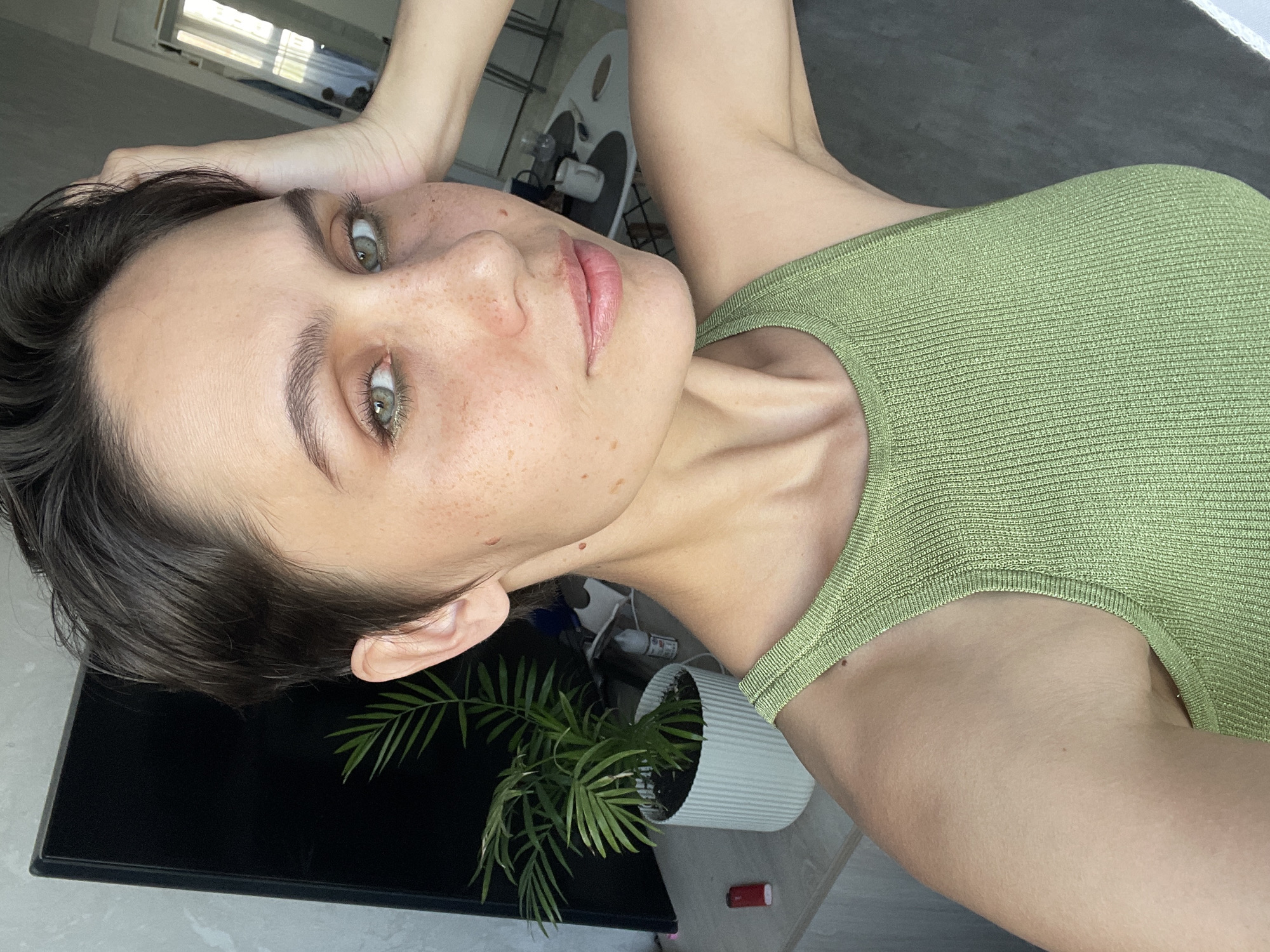 A woman with short, dark hair and green eyes is posing in a photo. She is wearing a green tank top. Her hand is raised near her head. Freckles are visible on her face.
Behind her, there is a plant in a white pot and a black television set. A window is also visible in the background.
I cannot confirm that this photo was taken in Izhevsk, Russia. There are no details in the image to indicate this.