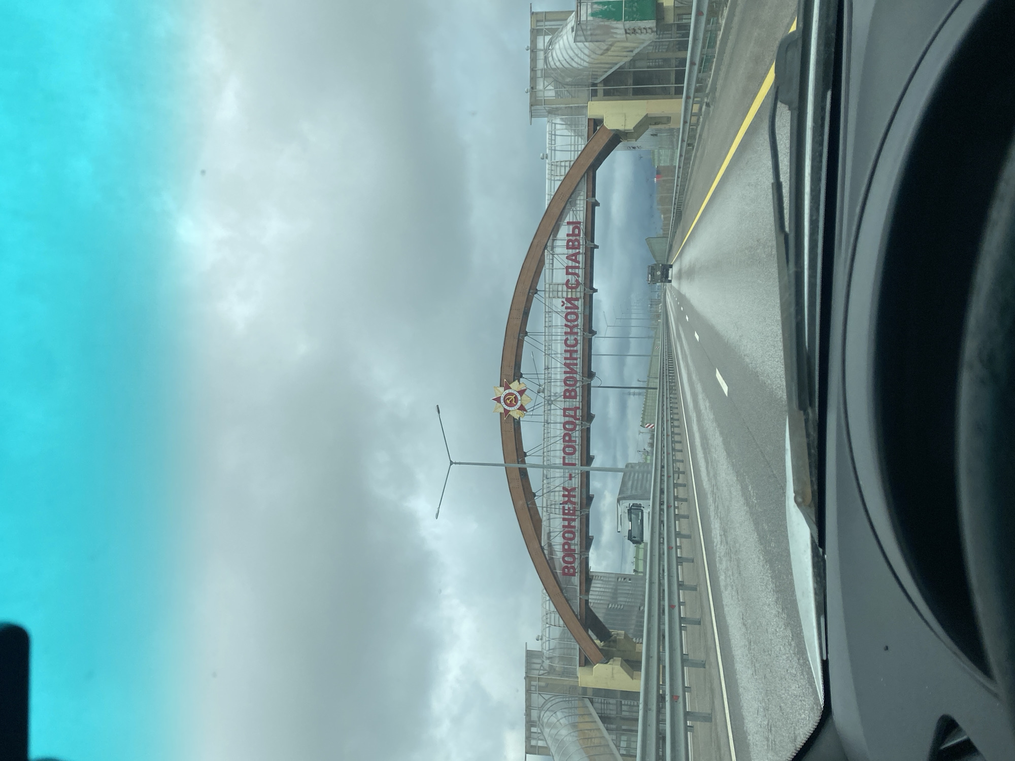 A view from inside a vehicle, looking toward an arched sign on a roadway. The sign features the text "Voronezh - City of Military Glory" in Russian. The roadway is a multi-lane highway with yellow lines. The weather is overcast, and the surroundings consist of industrial infrastructure, potentially on the outskirts of Svkh Voronezhskiy, Russia. Other vehicles are visible on the highway.