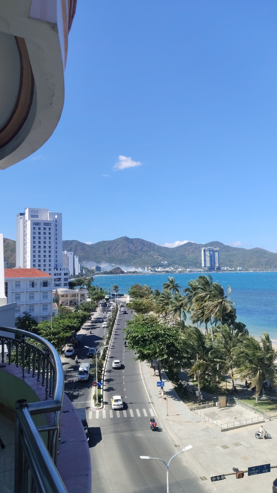 The image shows an elevated view in Bac Nha Trang Ward, Vietnam. A wide road runs from the foreground to the distant sea, populated with cars, scooters, and a three-wheeled vehicle. Buildings line the left side of the road, contrasting with the palm trees and beach on the right. Mountains are visible in the distance across the water. A hotel balcony occupies the left portion of the image, partially obscuring the view. The sky is clear and blue with a small cloud.