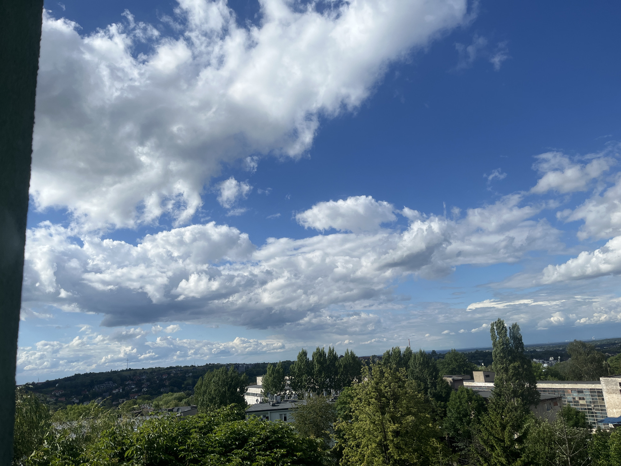 This is a medium shot, taken from a high vantage point, of a cloudy sky over a city. The sky is a vibrant blue with large, fluffy white clouds dominating the upper portion of the image. Below the sky, a densely wooded hillside stretches across the frame, dotted with houses and buildings. The trees are lush and green, with some buildings peeking through the foliage. In the foreground, the edge of a dark, vertical object is visible on the left side of the image, obscuring part of the scene. The overall impression is one of a clear, but cloudy, day in a suburban or semi-rural area within a city.