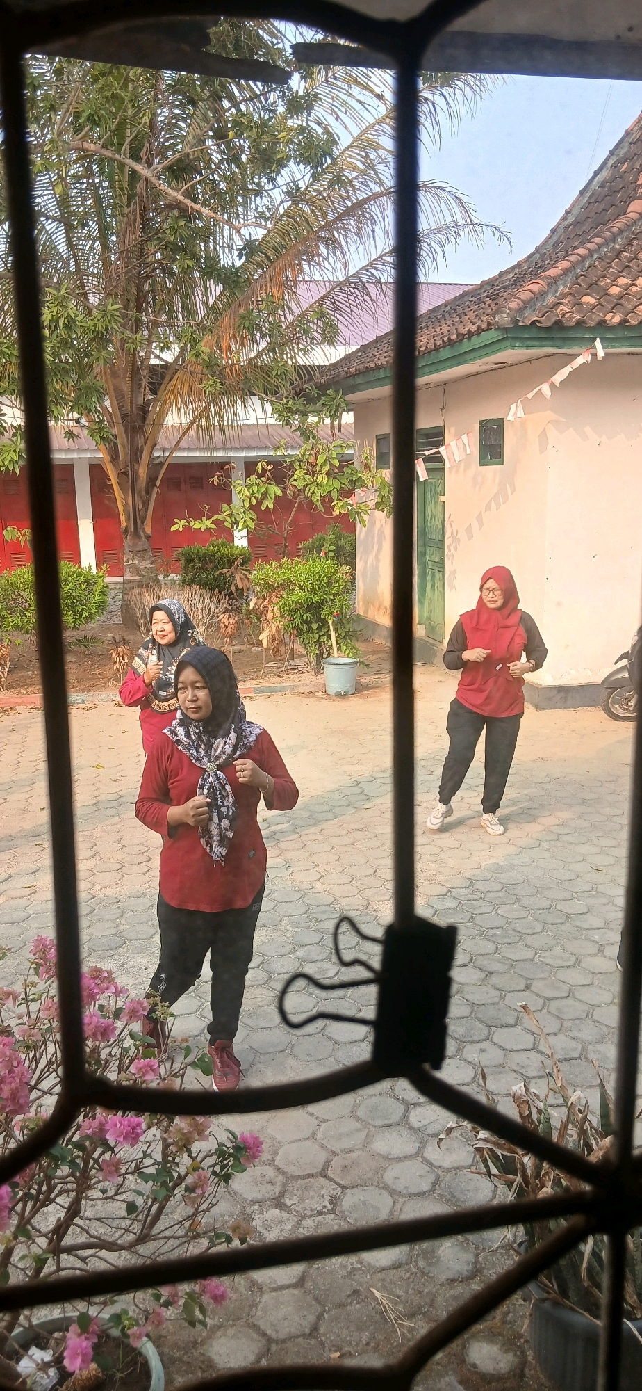 In an outdoor courtyard with hexagonal stone paving, three women are seen engaged in what appears to be physical activity. The woman in the foreground, facing the viewer, wears a maroon long-sleeved shirt, black pants, and a black and white patterned headscarf. She is in mid-stride, with her left leg forward. To her left and slightly behind, another woman, also wearing a maroon top and black pants, is jogging, looking towards the viewer. Her headscarf is black and white. Further back, to the right, a third woman in a red long-sleeved top and black pants, with a red headscarf, is also jogging. She is looking to her left.
The background features a building with a light-colored wall and a terracotta tiled roof. A green door is visible on the building. Lush green bushes and a palm tree are present, along with some potted plants. The scene suggests a warm, sunny day. The vantage point is from behind a metal grate or window bars, which partially obscure the view. The presence of what appears to be Indonesian flag decorations on the building implies a possible national holiday or event. The context suggests this location is likely within the city of Metro, Indonesia, given the architectural style and vegetation commonly found in the region.