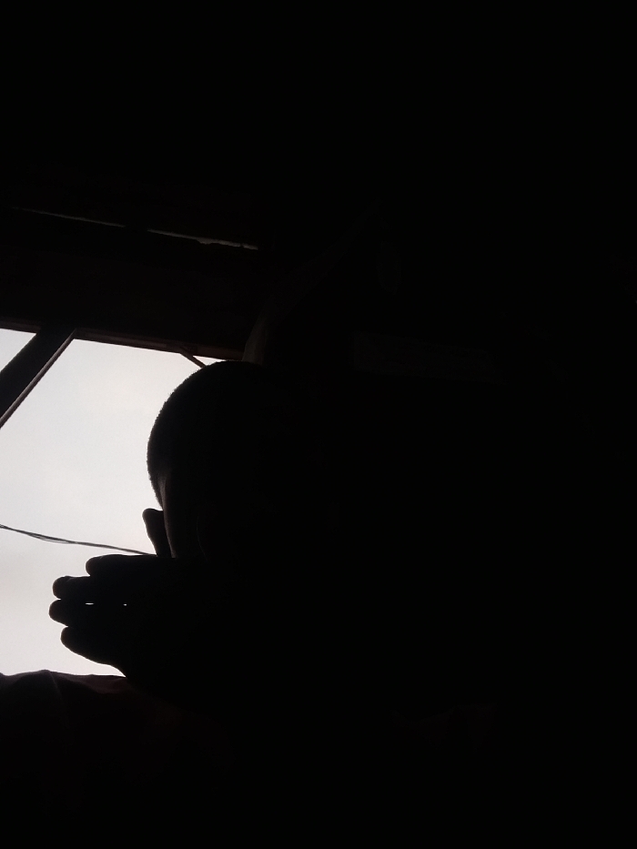 The scene is an interior view from Nteje, Nigeria, dominated by a dark silhouette of a human figure positioned against a brighter, overcast exterior visible through an opening on the left. The figure's head, exhibiting a short haircut, is discernible, and a right hand with distinct fingers is held near the lower face or neck area. The exterior light source illuminates a window frame or structural elements on the left, which appear dark. A thin, light-colored wire extends horizontally across the lower-left quadrant of the frame. The immediate foreground and upper-right portions of the image are in deep shadow, rendering further details of the interior environment obscure.